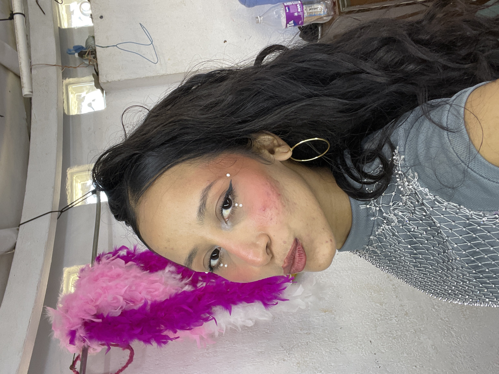 A young woman with dark, wavy hair is posed for a selfie in what appears to be a rustic indoor setting. She is wearing makeup, including blush, eyeliner, and small pearl-like dots under her eyes. She also has large gold hoop earrings and a unique, sparkly mesh top. Behind her, a pink and purple feather boa hangs on the wall. The background includes a white, textured wall with exposed pipes and electrical wires, and small square windows letting in natural light. A bottle of water is visible on a shelf in the background. The scene is indoors, likely in a residential or makeshift studio space.