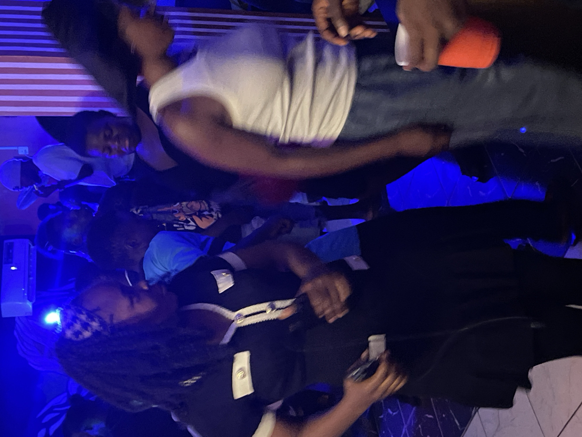 This image depicts a scene of revelry within a dimly lit, indoor venue in Durumi, Nigeria. The atmosphere is charged with blue lighting, reflecting off the dark, polished floor and the striped, vertical wall decor. In the foreground, a woman with long, dark hair adorned with a checkered headband is captured mid-dance, wearing a black dress with white trim and metallic buttons. Her right hand is raised, holding a dark object, possibly a phone. To her right, another woman, her face obscured, is also dancing, clad in a white tank top and blue pants. Her left hand is raised, holding a bright orange cup. In the background, other individuals are visible, some dancing, others observing the scene. One person in the mid-ground wears a blue shirt and appears to be dancing. The overall impression is one of a lively social gathering, possibly a party or a club night.
