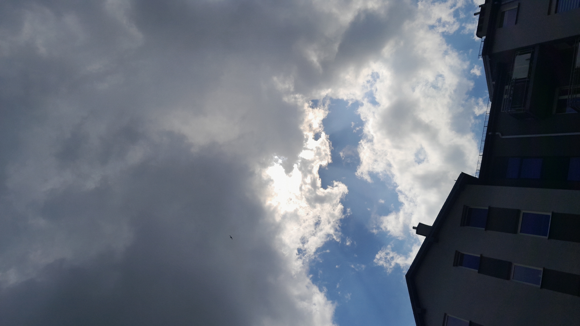 The image is an outdoor, low-angle shot of the sky with parts of two modern buildings visible on the right. The sky is a dramatic mix of dark, heavy clouds on the left and brighter, sunlit clouds with visible sunbeams on the right. A small bird is visible in flight against the dark clouds.
The time of day appears to be daytime, given the sunlight. The weather seems to be changing, with a storm potentially brewing or clearing, indicated by the contrast between the dark and bright clouds. The buildings are multi-story structures with visible windows. One building's roofline extends from the bottom right corner upwards, and another is partially visible at the top right. The architecture suggests an urban or suburban setting.
There is no discernible activity or event taking place beyond the natural atmospheric conditions and the flight of a bird. No text is visible in the image. The overall mood evoked is one of dynamic weather and perhaps a sense of awe at the power of nature.