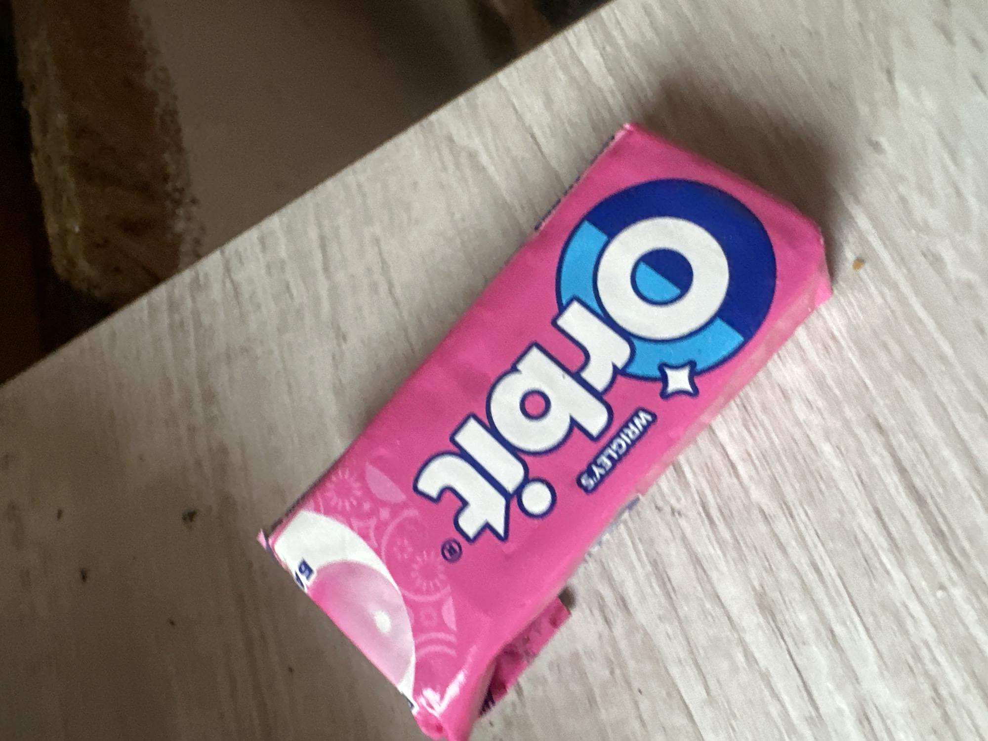 A package of Orbit chewing gum lies on a light-colored wooden surface. The gum package is pink with white and blue lettering. The brand name "Orbit" is prominently displayed in large white letters with blue outlines. Below "Orbit" is the word "WRIGLEY'S" in smaller white letters. The background appears to be a wooden table or countertop. The lighting suggests an indoor environment, possibly a home or office. There are no people or animals visible in the image. The scene is static and appears to be a still life. The location in Odesa, Ukraine is not discernible from the image.
