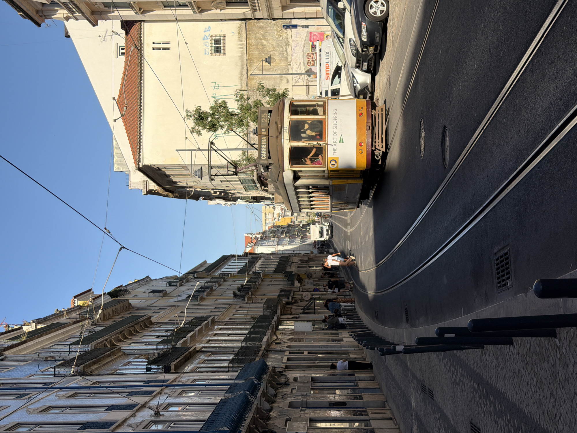A tram travels down a narrow, steep street in Lisbon, Portugal. The tram is yellow and white with "THE ART OF SHOPPING" written on its side. Several people are visible inside the tram. The street is lined with tall, historic buildings with many windows and balconies. The sky is clear and blue, suggesting a sunny day. The sun casts long shadows, indicating it is likely late afternoon. Several people are also visible on the sidewalks, some sitting on steps and others walking. Tram tracks run down the center of the street. A black car and a white van are parked or driving further up the street. The overall scene depicts a bustling urban environment in a European city.
