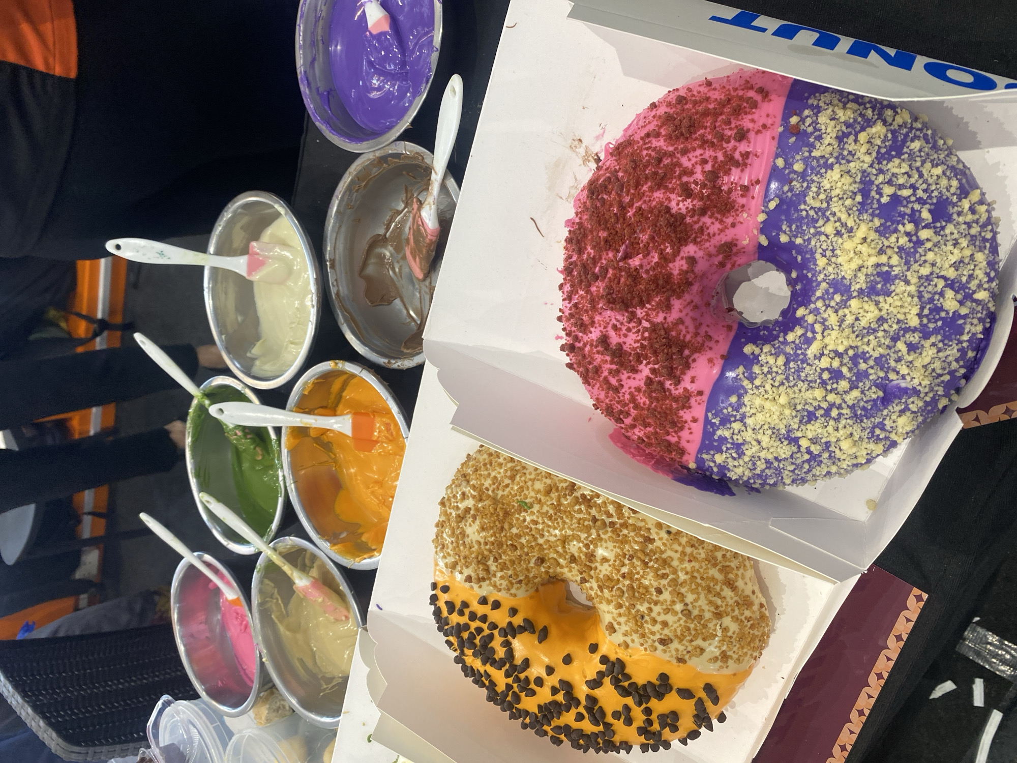 The image depicts a close-up, eye-level view of two donuts in white cardboard boxes, presented at a stall or event. In the background, several metal bowls are visible, each containing a different colored frosting and a spatula: pink, green, tan, orange, brown, and purple. These bowls appear to be set up for decorating donuts. The donuts themselves are prominently displayed. The donut on the left is half-covered in orange frosting with chocolate chips and half in tan frosting with small, crunchy sprinkles. The donut on the right is half-covered in pink frosting with red crumble topping and half in purple frosting with yellow crumble topping. The overall scene suggests a dessert-making or tasting event, potentially a pop-up shop or a booth at a fair. The lighting is bright and focused on the donuts. The backdrop is blurred but suggests an indoor setting with hints of people and furniture. The specific location of Davao City, Philippines is not discernible from the visual information provided in the image.
