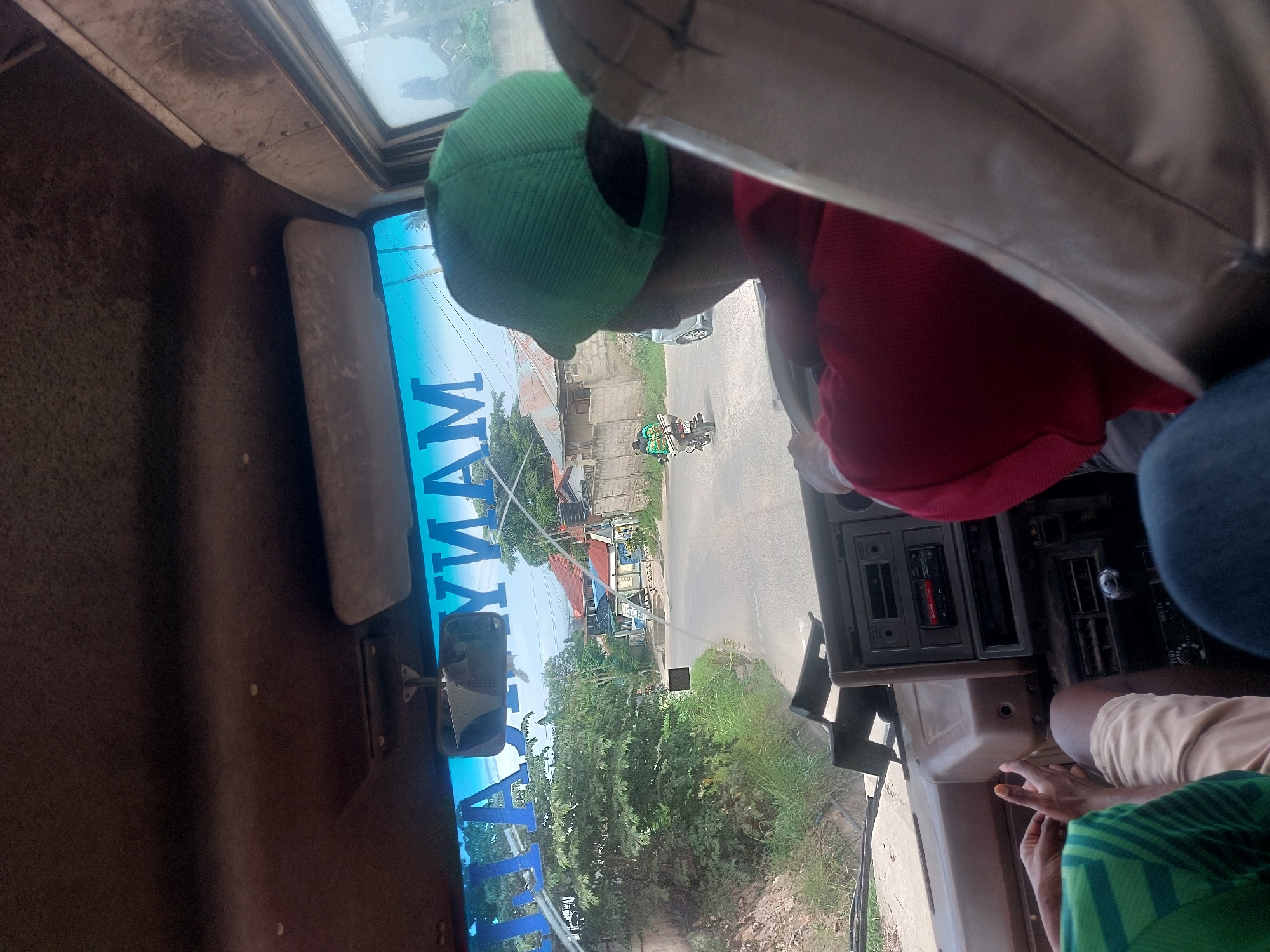 The image, taken from inside a vehicle, shows a view of a street in what is likely a residential area of Dar es-Salaam, Tanzania. The name "Mamyam" is visible, potentially indicating a business name or route identifier. A person wearing a green cap and red shirt is in the front seat. Through the windshield, one can see buildings, vegetation, and a road with a motorcycle and possibly a car. The vehicle's dashboard is visible, along with other passengers.