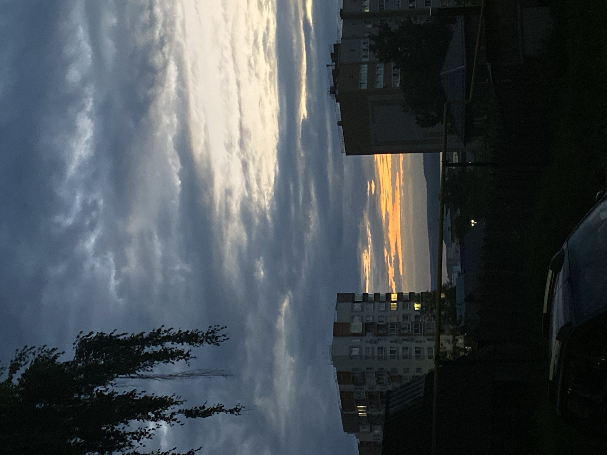 This image depicts a twilight scene in the city of Votkinsk, Russia, likely taken from an elevated position looking towards the west. The sky is dominated by dramatic, dark clouds with streaks of orange and yellow light breaking through near the horizon, indicating the setting sun. In the mid-ground, two apartment buildings are visible. The building on the left is lighter in color with orange accents and illuminated windows. The building on the right is darker and appears to be a residential block with many windows. Between and below these buildings, a darker silhouette of trees, smaller houses, and possibly a body of water can be seen in the distance. In the foreground, the dark shape of a car's roof and a section of a dark structure, possibly a fence or another building, are visible. A branch of a tree with long, thin leaves hangs from the upper left corner of the frame, partially obscuring the sky. The overall atmosphere is somber and peaceful.