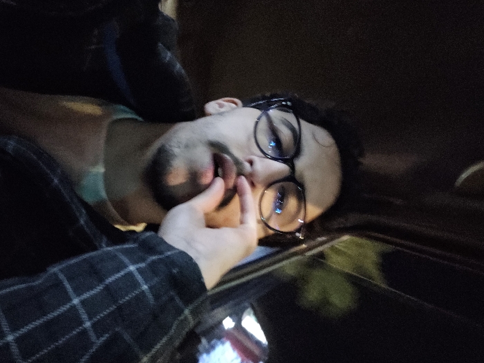 A person wearing glasses lies on their side, a hand touching their mouth. The individual has dark hair and a light-colored complexion. They are wearing a light blue undershirt and a dark plaid shirt. The background is mostly dark. A window with a reflection is in the foreground, and some lights are visible through the window. The image appears to be taken in a car at night.