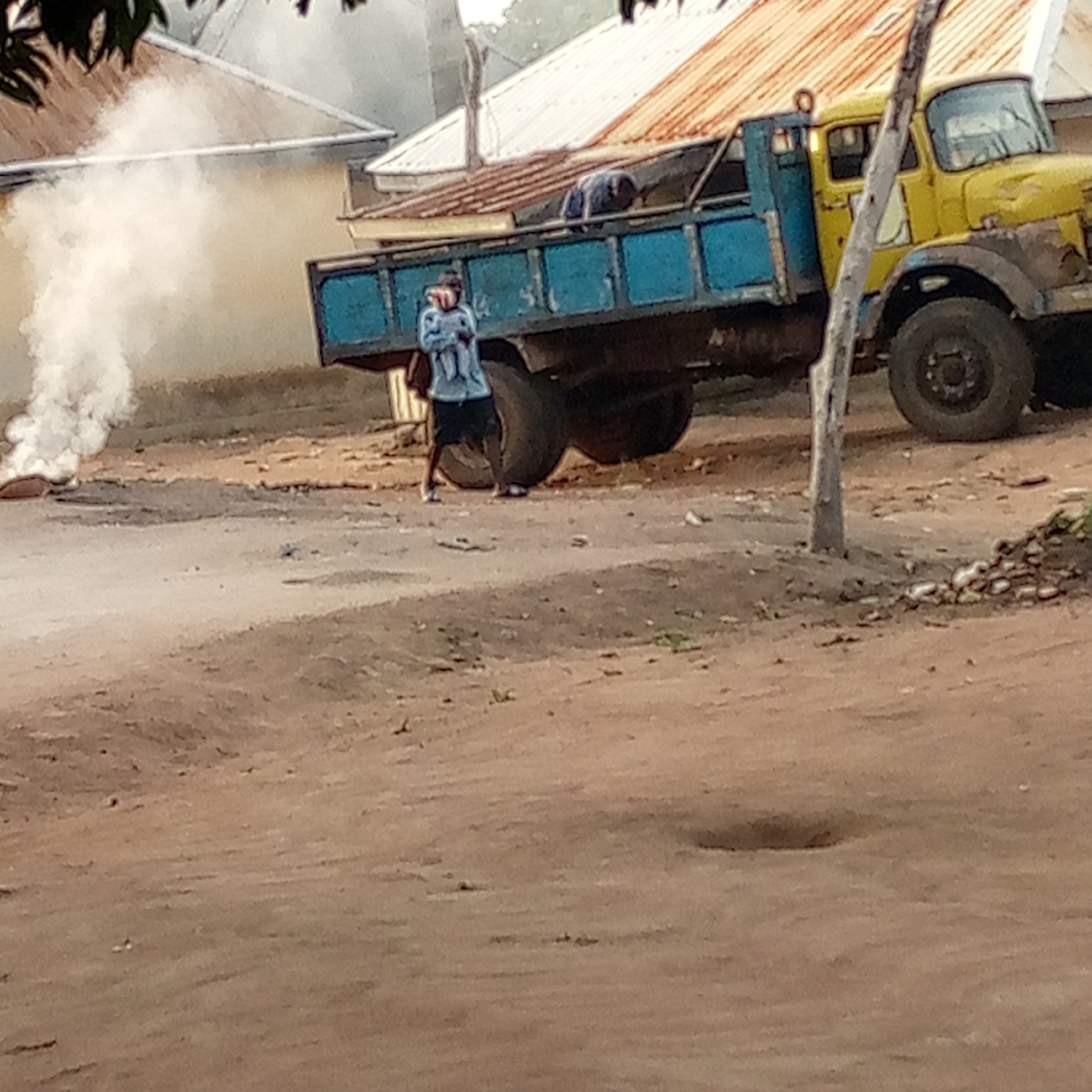 The image shows a street scene in what appears to be Mararaba, Nigeria. A yellow and blue truck is parked on a dirt road. A person wearing a light blue shirt and dark shorts is standing near the truck's rear tires. Another person is in the back of the truck, near the front. There is a plume of white smoke rising near the left edge of the frame, possibly from a fire. The road is dirt and appears to be unpaved. There are buildings visible in the background with what appears to be corrugated metal roofing. A thin tree trunk is to the right of the truck.