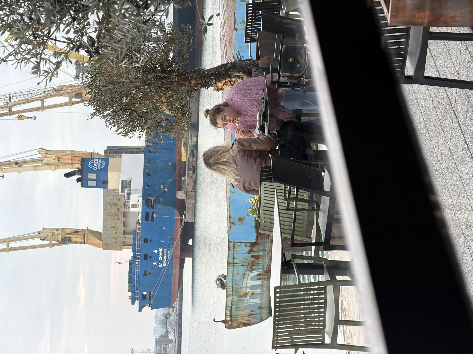 The image captures a scene at a waterfront location in Burgas, Bulgaria. Two women are seated at an outdoor table. One is wearing a pink sweater and eyeglasses, eating pasta with a fork. The other woman is wearing a tan jacket. There is a large blue cargo ship docked in the background with cranes visible. The ship is identified as the "DA CHANG" with its port of registry as "SINGAPORE." The outdoor seating area includes metal chairs and tables. The photo is taken from a partially obscured perspective, with elements of a structure (possibly a window frame) on the right side.