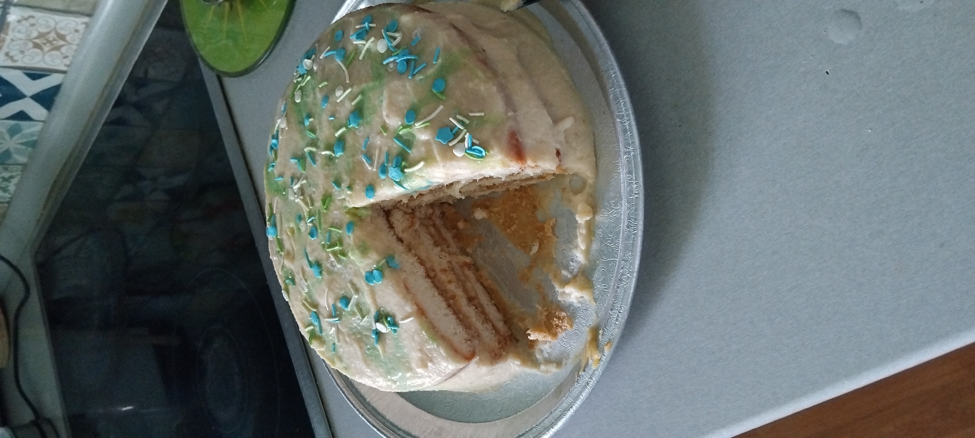 A cut cake with multiple layers sits on a silver platter on a light gray surface. The cake is frosted with a light-colored icing and decorated with green and blue sprinkles. A slice has been removed from the cake, revealing its layered interior and spilling some crumbs onto the platter. The background shows a kitchen counter with a black stovetop and a colorful tiled backsplash. The image's perspective suggests it was taken from a slightly elevated angle. There is no indication of the city of Chekhov, Russia, in the image.