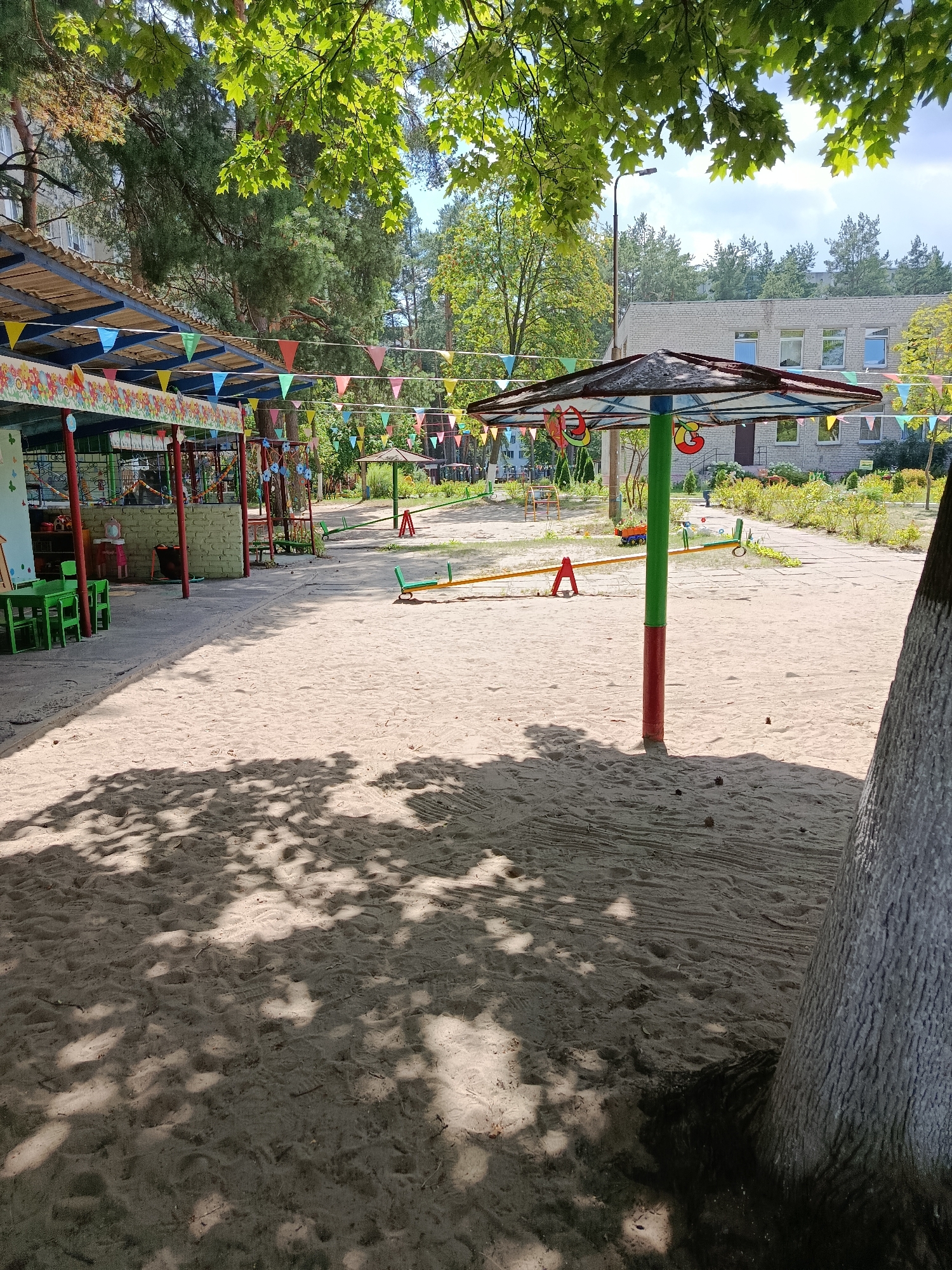 The image depicts a sandy playground area with various play structures. In the foreground, a large tree trunk is visible on the right side, with dappled sunlight casting shadows across the sand. A colorful canopy with pennant banners stretches across the left side of the image, sheltering a section of the playground with tables and chairs. In the middle ground, a seesaw is set up on the sand, and further back, a smaller shaded structure with a colorful top is visible. Beyond the playground, a building with windows and a brick facade is present, surrounded by trees and greenery. The scene appears to be a typical outdoor playground, likely part of a school or kindergarten, located in the city of Svietlahorsk, Belarus. There are no people visible in the image.