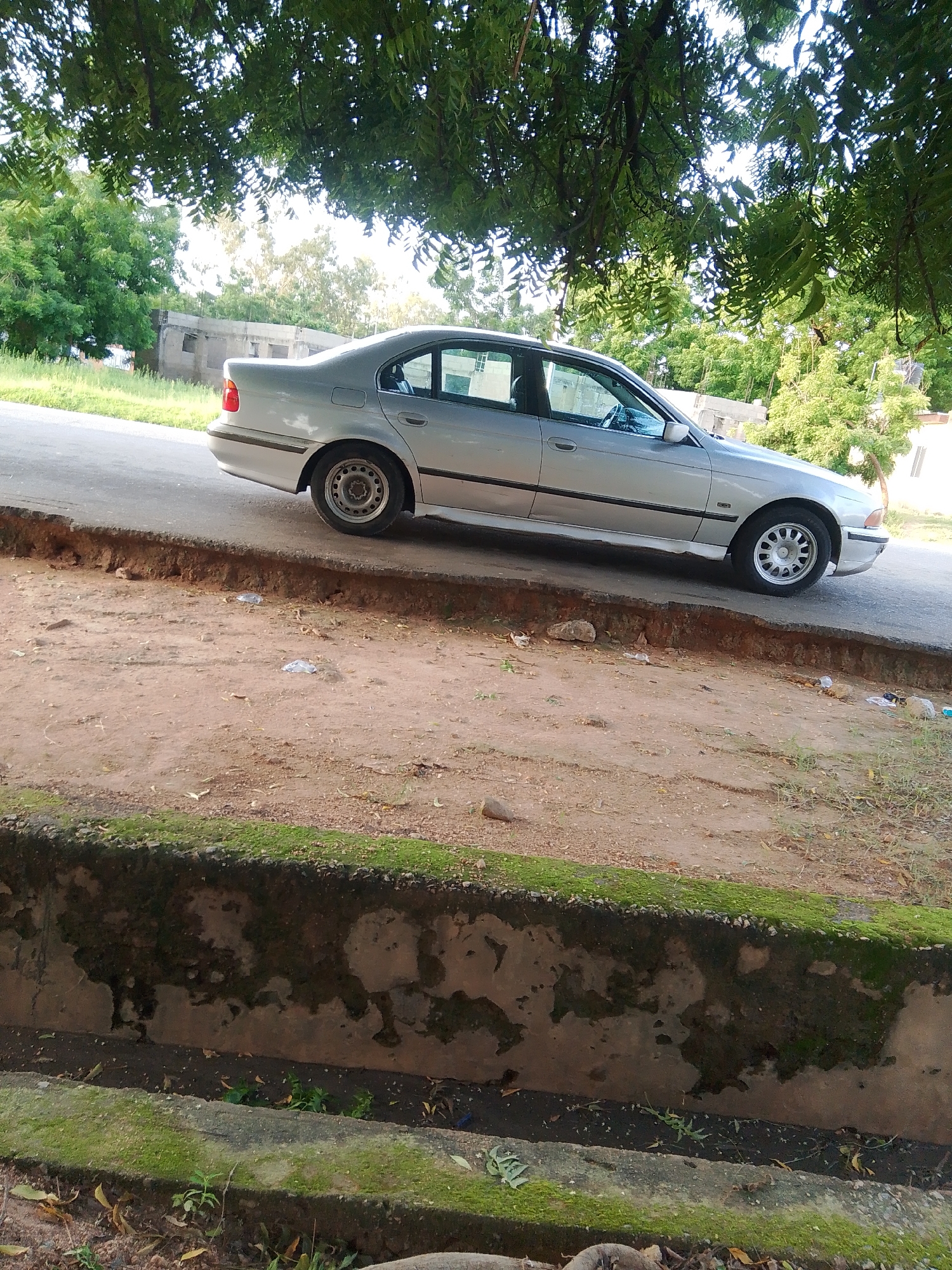 A silver sedan is parked on the side of a paved road. The car is partially under the shade of a large tree with green leaves. The road has a rough, uneven edge, and the ground next to it is a dirt path with some scattered debris. In the background, there are more trees and some buildings, one of which appears to be under construction or in disrepair. The overall setting suggests an outdoor, possibly semi-rural or older urban environment. The lighting indicates it is daytime, with sunlight filtering through the tree canopy. There is no visible text in the image. The scene appears still, with no people or active events taking place. The mossy growth on the concrete structure in the foreground suggests a humid or damp environment.