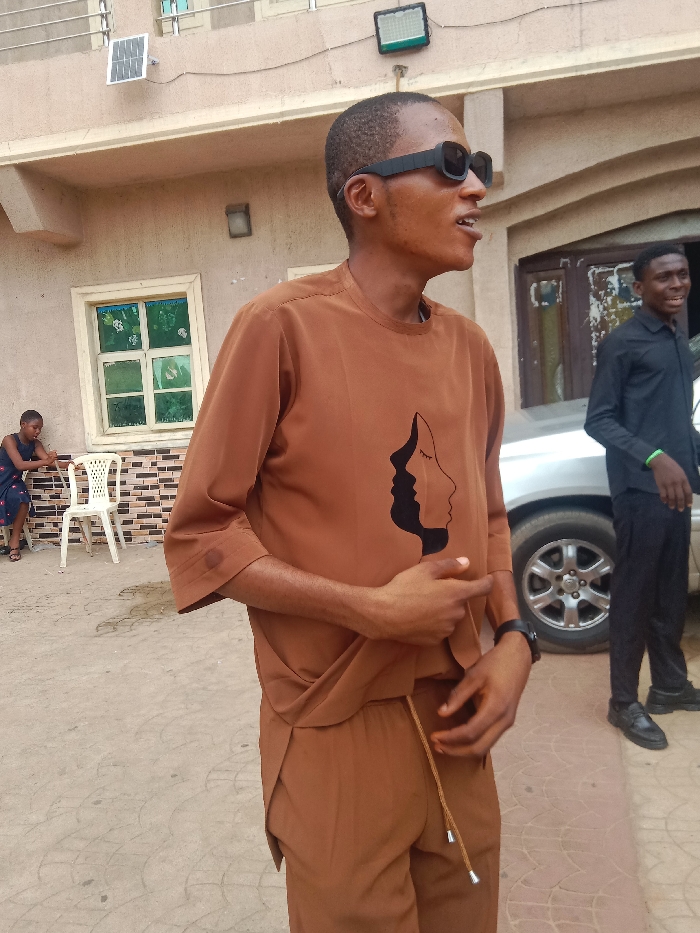 This image depicts an outdoor daytime scene in Onitsha, Nigeria, featuring three individuals and a building facade. In the foreground, an adult male with dark skin and short hair wears black rectangular sunglasses and a brown two-piece outfit, which includes a tunic with a black abstract design of two faces on the chest and matching trousers with a visible drawstring. He is positioned facing right, with his mouth slightly open, and his right hand is held near his stomach. A black wristwatch is present on his left wrist. To his immediate left and slightly behind, a young female with dark skin is seated on a low surface, partially obscured by a white plastic chair. She wears a dark patterned dress. Behind her is a light beige building with a white-framed window featuring green-tinted glass panes. Above the window, a small solar panel and a dark-colored floodlight are affixed to the building. To the right, a second adult male with dark skin, dressed in a black long-sleeved collared shirt and black trousers, stands next to a silver sedan, partially visible behind him. He wears a light green wristband on his right wrist and is smiling, looking towards the central figure. The ground throughout the scene is paved with light-colored interlocking patterns.