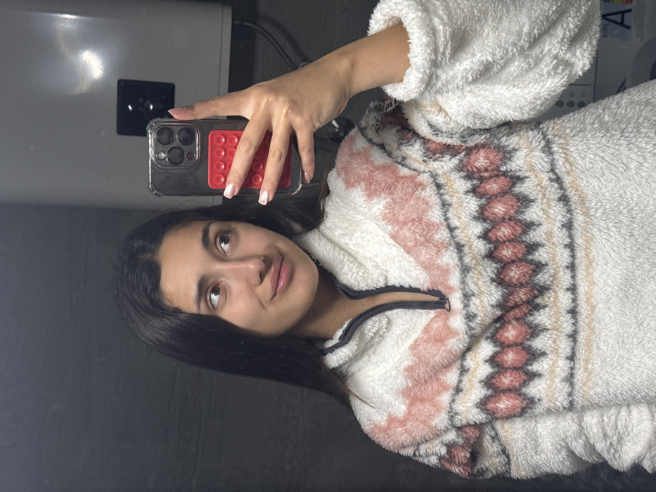 A young woman poses for a selfie, holding her phone with a clear case and a red pop socket. Her hair is dark and long. She wears a fluffy white sweater with a zip-up neckline and a patterned yoke in shades of pink, brown, and beige. The background is a plain gray wall, with a white appliance visible in the upper left corner.