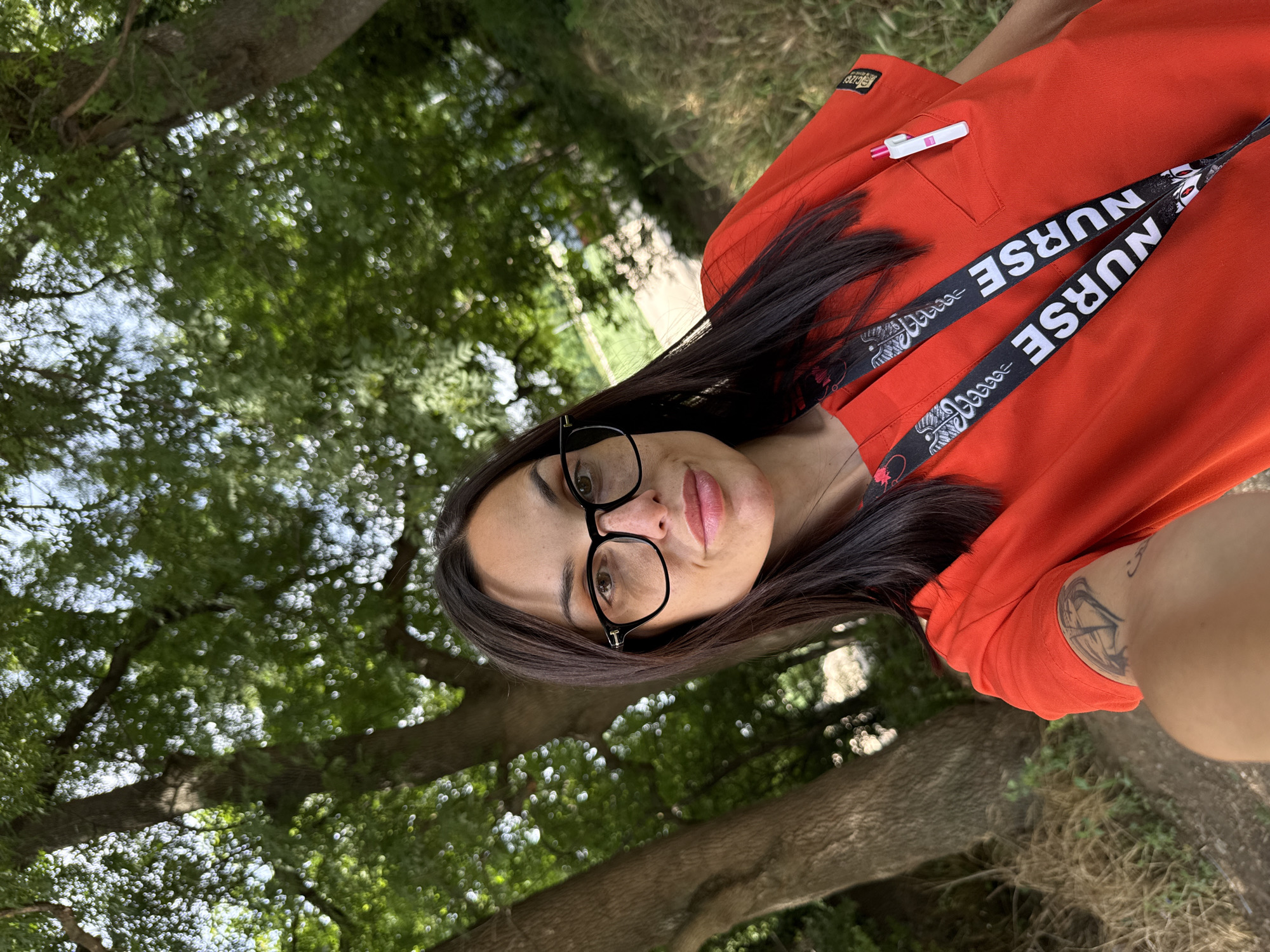 A selfie-style portrait captures a woman outdoors, looking directly at the camera. The image is rotated 90 degrees counter-clockwise. She has long, dark hair, dark-rimmed glasses, and appears to be wearing light pink lipstick. She is dressed in a bright orange short-sleeved scrub top, which features a small emblem on the right chest pocket and a white pen with a pink clip in the left chest pocket. Around her neck is a black lanyard with the word "NURSE" printed repeatedly in white, along with medical symbols, possibly a Caduceus or staff of Asclepius. A tattoo is visible on her left arm.
The background consists of lush green trees and foliage, blurred to emphasize the subject, suggesting a park or natural outdoor setting, possibly in Pleven, Bulgaria. The lighting indicates it is daytime. The overall mood is casual and natural.