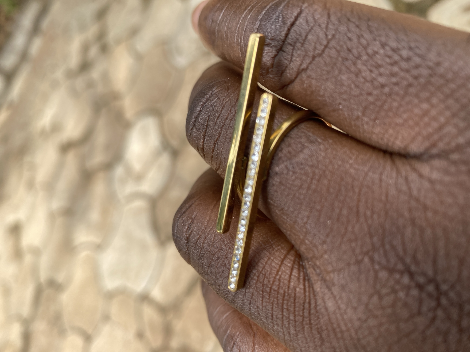 A close-up captures a person's brown hand, adorned with a gold-colored ring. The ring features a horizontal bar on top and a vertical bar accented with small, bright stones. The background is an out-of-focus pattern of light tan and brown shapes. The location is assumed to be Maigizo, Nigeria.