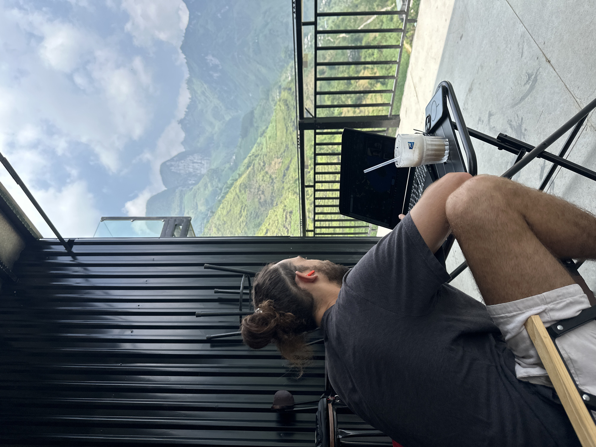 A man with long hair tied in a bun sits on a chair outdoors, working on a laptop. A drink with a straw sits on a table next to the laptop. In the background is a mountainous landscape, likely in Mèo Vạc Commune, Vietnam. The man is wearing a gray shirt and light-colored shorts. The scene appears to be a balcony or terrace with a black metal railing and a black corrugated metal wall.