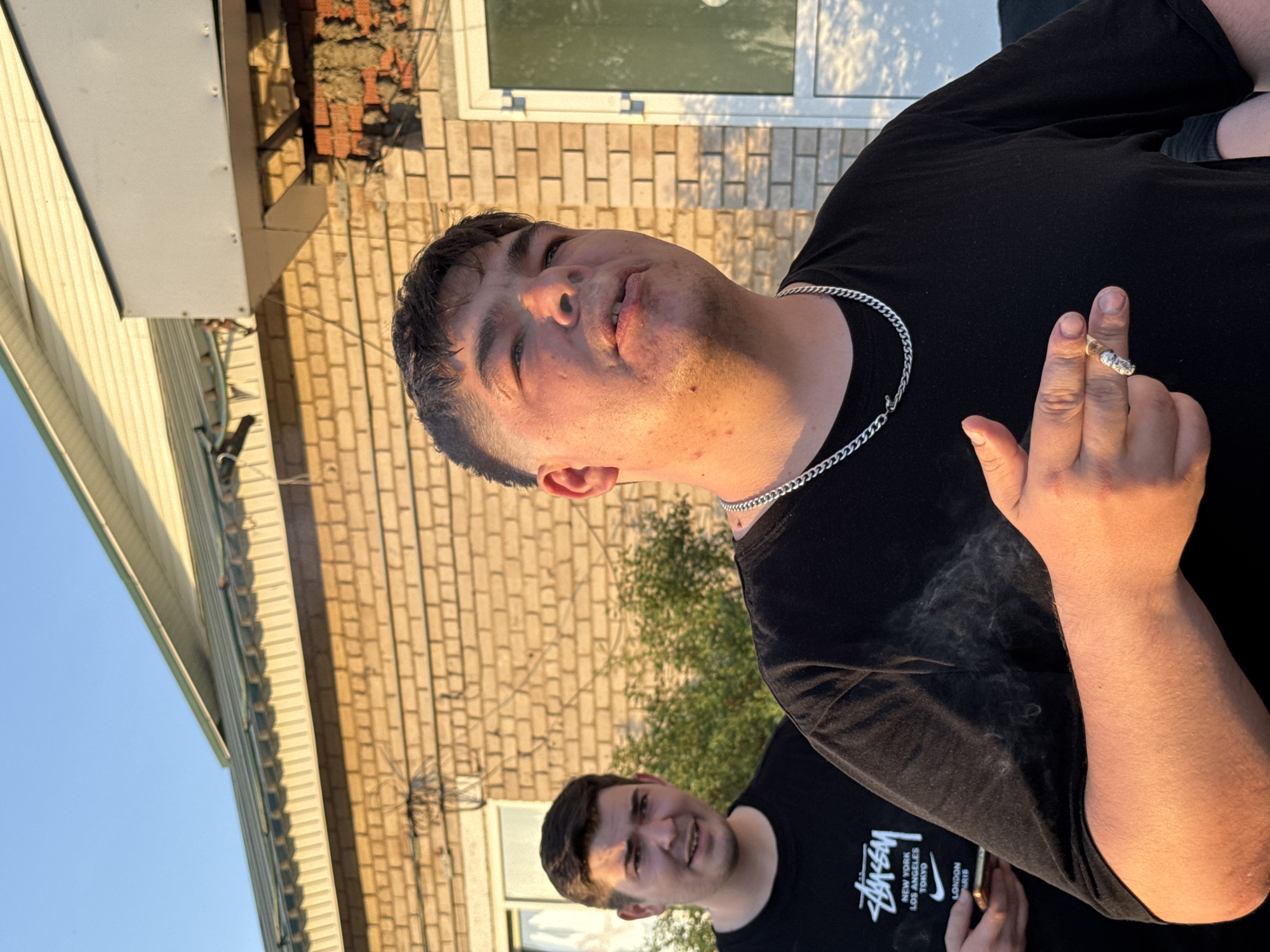 A photograph shows two men in an outdoor setting. The main subject in the foreground is holding a lit cigarette, from which smoke is emanating, and has a silver chain around his neck. He is wearing a black t-shirt. Partially visible behind him and to the left is another man, also wearing a black t-shirt with a white Stussy logo. In the background, there is a brick building with a window. To the left of the brick building is a light yellow structure. The location is potentially in the city of Кемуль, Russia.