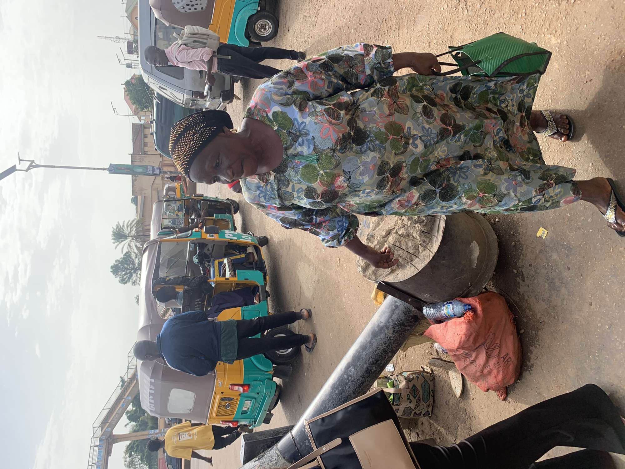 A woman in a colorful floral dress and a black patterned headscarf walks on a dirt road in Gura Top, Nigeria, in front of a beige and green auto-rickshaw. She is carrying a green woven bag in her right hand and wearing beaded sandals. Behind her, a man in a pink shirt and backpack stands near a silver van. Other auto-rickshaws and pedestrians are visible in the background. The scene appears to be a busy street or marketplace under a partly cloudy sky.