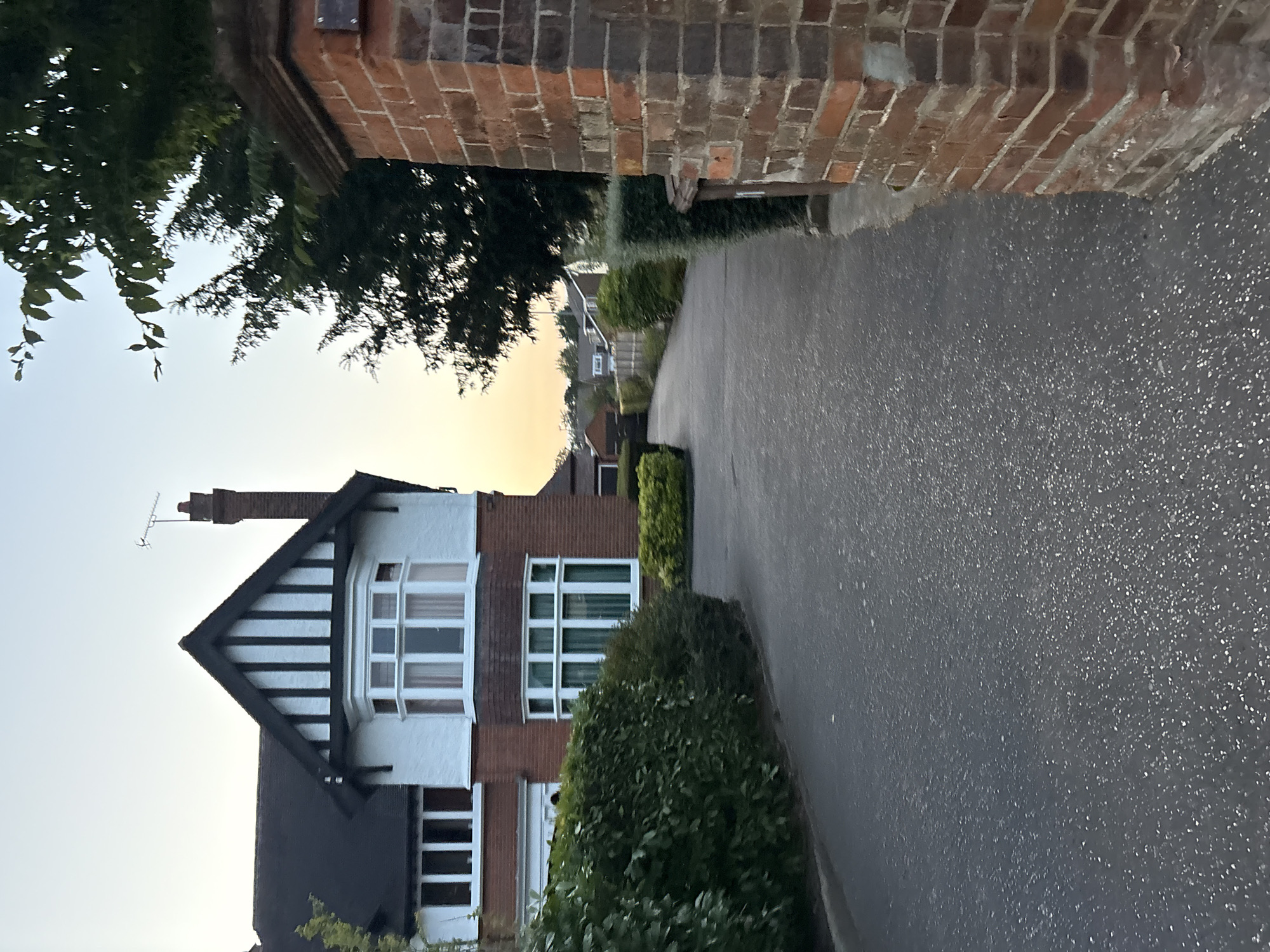 The image captures a residential street scene in Chelmsford, United Kingdom, likely at dusk due to the soft, warm light in the sky. A large, detached house with a distinctive black and white timber-framed gable is prominently featured on the left. The house has red brick lower walls and white upper walls, with multiple large windows, including a bay window. A paved driveway or path leads away from the house, curving to the right. On the right side of the image, a brick pillar marks the entrance to a property, constructed from red bricks. Trees and manicured hedges line the driveway, creating a sense of privacy. The sky is clear with a gradient of pale yellow to blue, typical of twilight. The asphalt surface of the driveway is visible in the foreground, speckled with reflective particles. No people or vehicles are present in the scene.