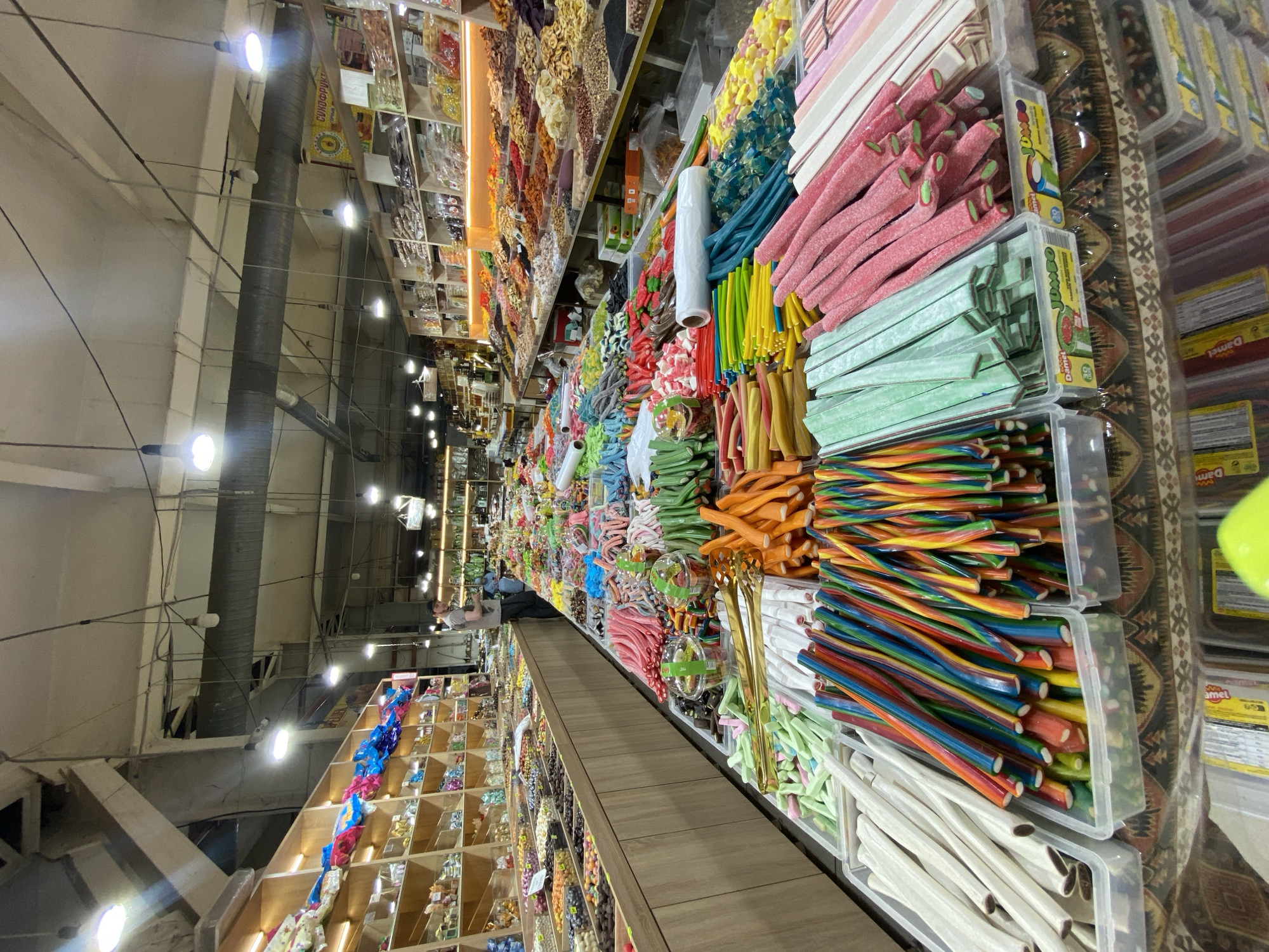 This media file captures a vibrant candy store scene in Makhachkala, Russia. The camera is positioned to offer a sweeping view of aisles filled with an immense variety of sweets, predominantly gummies and licorice. The primary subjects are the colorful candies themselves, displayed in clear plastic bins and on shelves, creating a visually stimulating and abundant selection. Among the candies are various shapes and colors, including long, thin licorice sticks in rainbow hues, chunky pink and green gummy strips, and smaller, brightly colored gummy candies.
In the background, the setting appears to be a well-lit indoor market or a large retail space with a high ceiling and exposed industrial elements like pipes and electrical conduits. Wooden shelves are visible, stocked with packaged goods and more bulk candies. A few people are present, including a man in the background wearing a light-colored t-shirt, possibly a shopkeeper or customer, interacting with the merchandise. Another person, partially obscured, is seen in the far background, suggesting the store is open and has customers.
The overall impression is one of abundance and sensory appeal, with the bright colors of the candy dominating the scene. There are no clear indicators of the time of day or weather. The atmosphere appears to be that of a bustling marketplace. Visible text includes partial labels on candy packaging, such as "Jumbo" and "Damol," and some Cyrillic text on labels within the shelves, though it is mostly illegible. The rich display of confectionery suggests a popular spot for sweets.