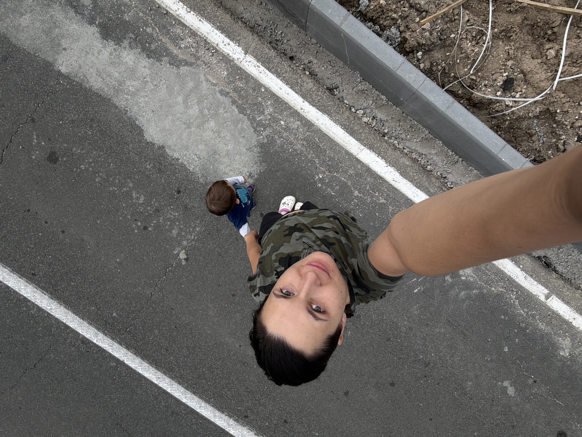 The image is a high-angle selfie taken outdoors, showing two subjects on an asphalt surface marked with white lines. One subject, in the foreground and center, is a person looking up at the camera, wearing a camouflage-patterned shirt. Their face is visible, and they appear to be taking the photo with their right arm outstretched.
To the left and slightly behind the first subject is a young child, facing away from the camera and looking downwards. The child is wearing a blue shirt with a graphic design and dark pants.
The scene is a paved area, possibly a road or walkway, with a concrete curb and dirt/gravel on the right side. The lighting suggests it is daytime, but the weather is overcast or diffused. There is no visible text in the image. The overall impression is of a casual outdoor moment between an adult and a child.