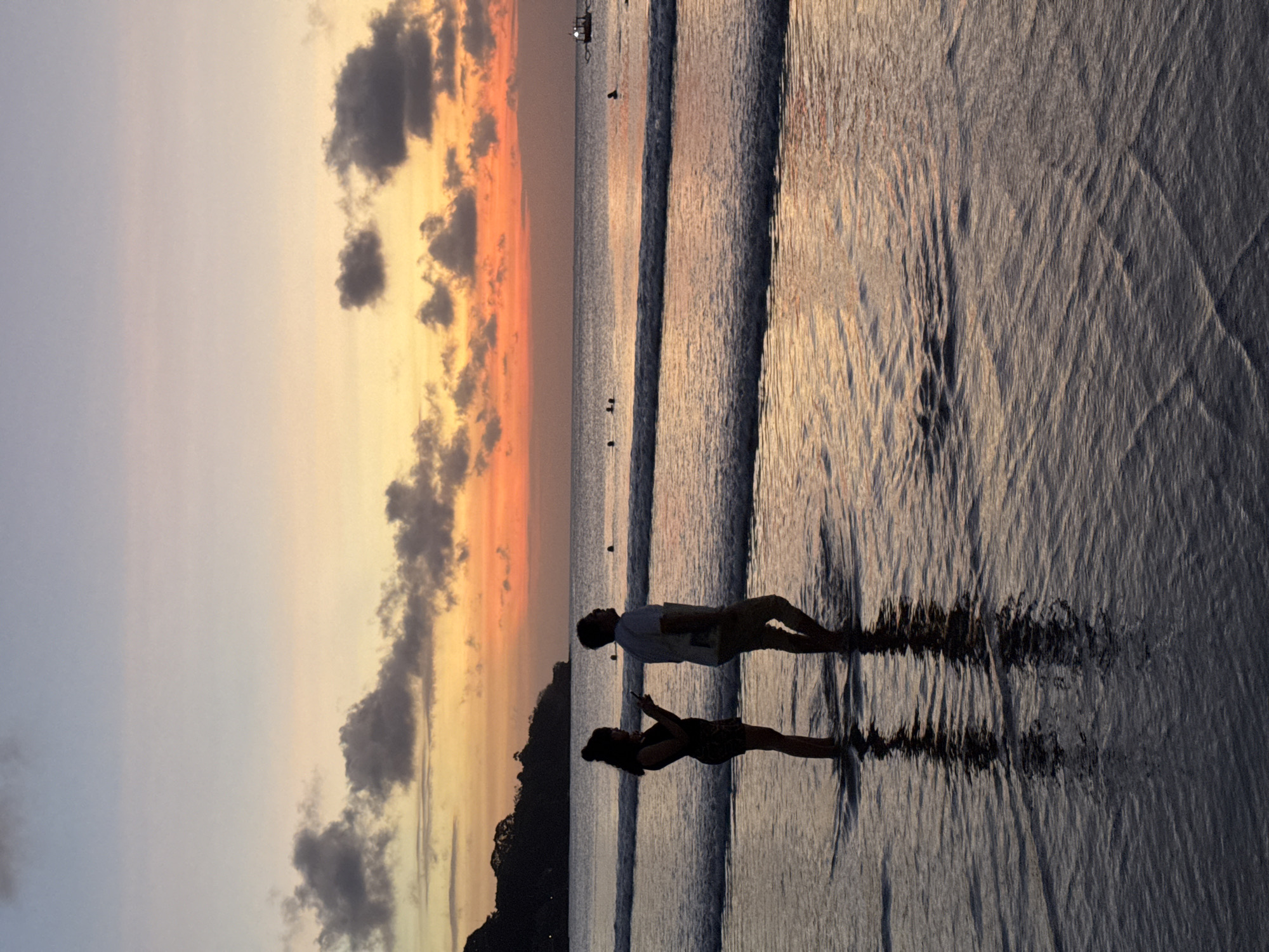 The image depicts a serene sunset scene at a beach in Kuta Selatan, Indonesia. The sky is a gradient of soft hues, transitioning from a pale lilac on the left to warm oranges and yellows as it nears the horizon, with scattered dark clouds adding texture. Silhouetted against this vibrant sky are two people standing in the shallow water. A man, wearing a light-colored short-sleeved shirt and shorts, faces a woman who is looking down at her phone. The woman is dressed in dark clothing and her hair is tied up. Their reflections are clearly visible on the wet sand and rippling water, creating a sense of symmetry and calm. Gentle waves are breaking on the shore, their foam creating delicate patterns on the wet sand. In the distance, the ocean stretches out, with a few small boats visible on the horizon, hinting at a calm sea. A dark, silhouetted landmass, possibly a hill or a section of coastline, is visible on the far left. The overall mood is peaceful and romantic, evoking a sense of quiet enjoyment of the natural beauty of the sunset. The image appears to have been taken during the golden hour, just before sunset. No text is visible in the image.