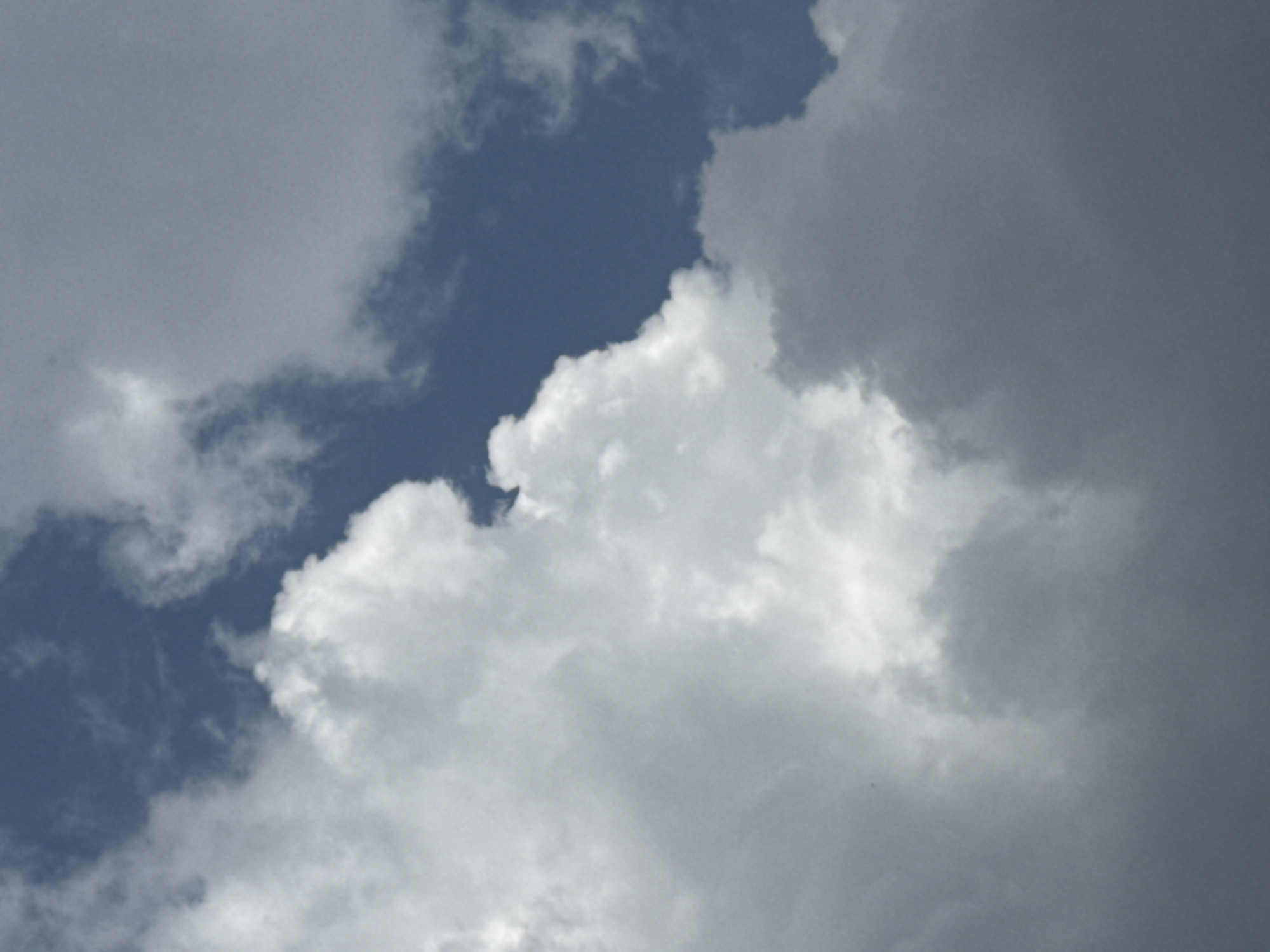 This is a medium-shot, eye-level, vertical, natural light photograph of clouds in the sky over Đa Lộc, Vietnam. The sky is a mix of blue and white, with large, fluffy white clouds dominating the left side of the frame. The clouds are illuminated from above, creating bright highlights and soft shadows. The blue sky is visible in patches through the clouds, particularly in the upper right corner. The overall impression is one of a bright, airy day.
