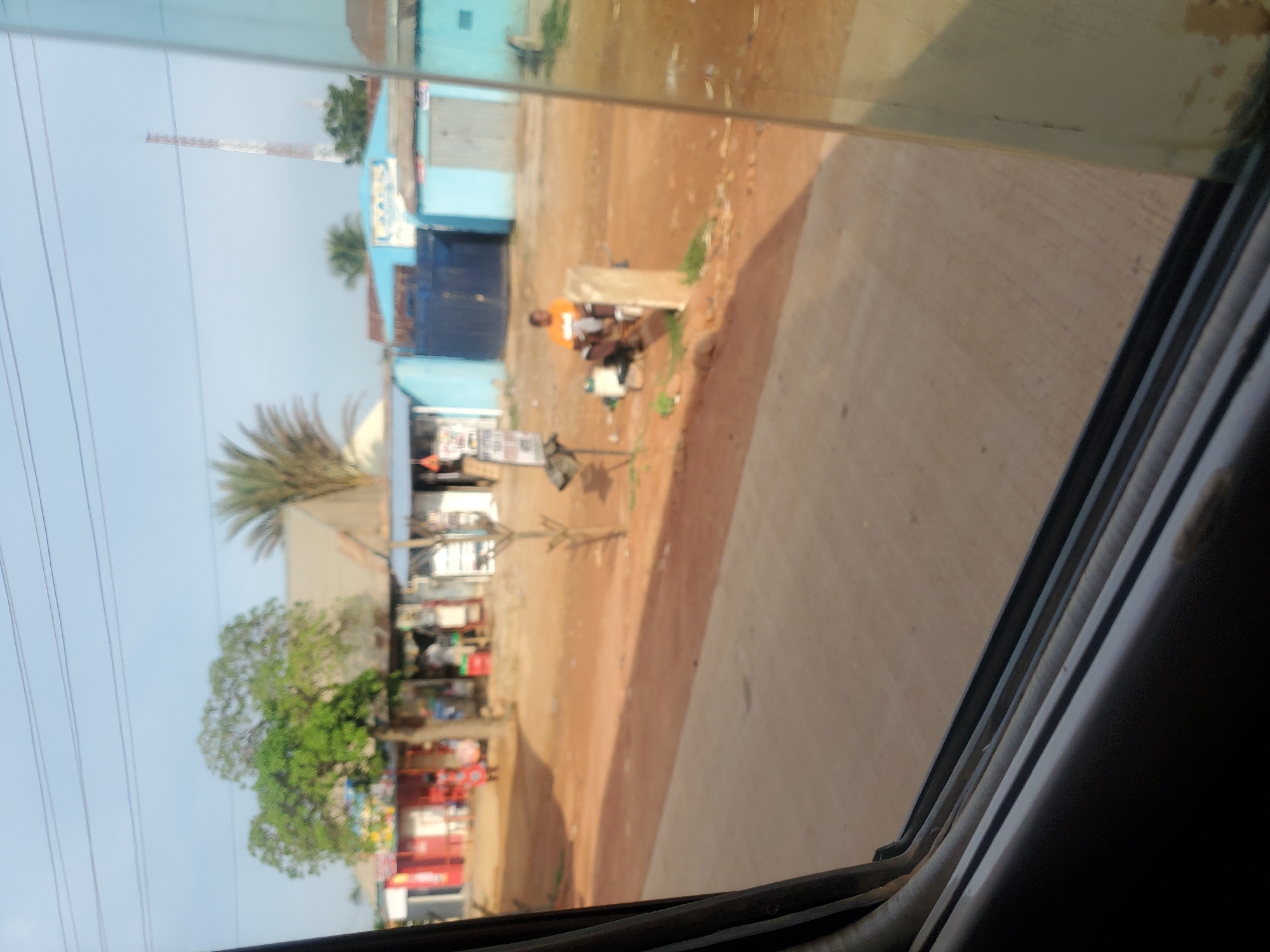 The image depicts a street scene in Kakau, Nigeria. In the foreground, a portion of a vehicle window is visible, framing the view of the surroundings. The street itself is made of dirt and gravel, with a paved road running through it. Several small businesses and residential structures line the street, some painted in bright colors. A few people are visible, including a person in an orange shirt sitting near a post and a woman in a patterned dress walking in front of a building. There are also trees and a telecommunications tower in the distance. The overall impression is that of a typical day in a bustling Nigerian town.