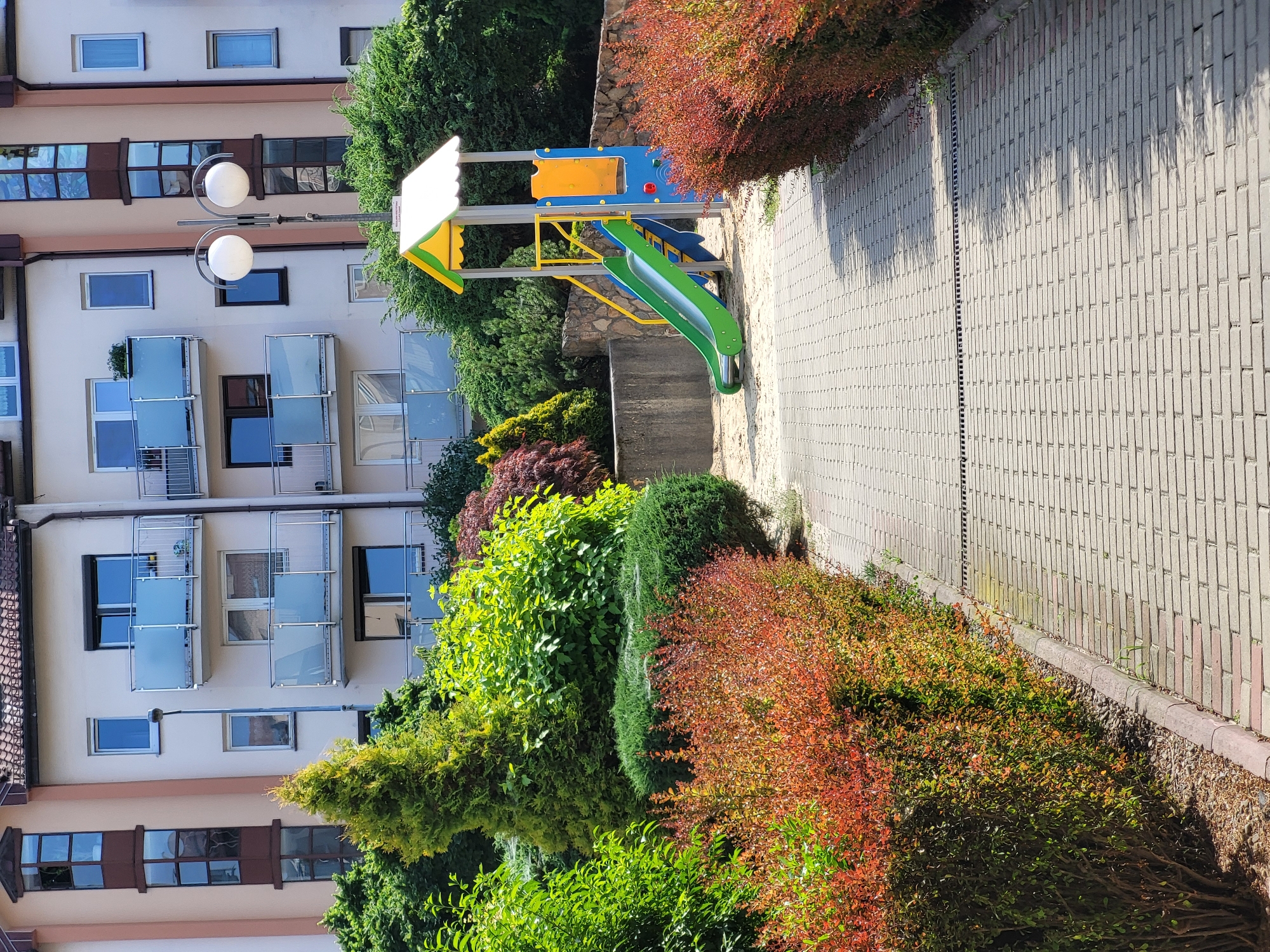 This is a medium shot, taken from a slightly elevated angle, of a residential courtyard with a playground. The scene is oriented vertically, with the top of the image pointing downwards. On the left, a multi-story apartment building is visible, featuring beige and cream colored walls, multiple windows, and balconies with glass railings. The windows are a mix of dark-framed and lighter-framed units. A street lamp with two spherical white lights is positioned in front of the building.
To the right of the building, a small playground structure with a bright yellow and green canopy and a blue and yellow panel is situated. A green slide extends from the structure down towards a sandy area. The playground is surrounded by lush greenery, including bushes with red and green foliage, and manicured hedges.
The foreground is dominated by a paved walkway made of light gray rectangular bricks. The walkway has a drainage grate running along its edge next to the bushes. Sunlight casts shadows from the foliage onto the brick path, suggesting it is daytime. The overall atmosphere is bright and sunny, conveying a sense of normalcy and everyday life in a residential area. No people are visible in the image. There is no visible text.