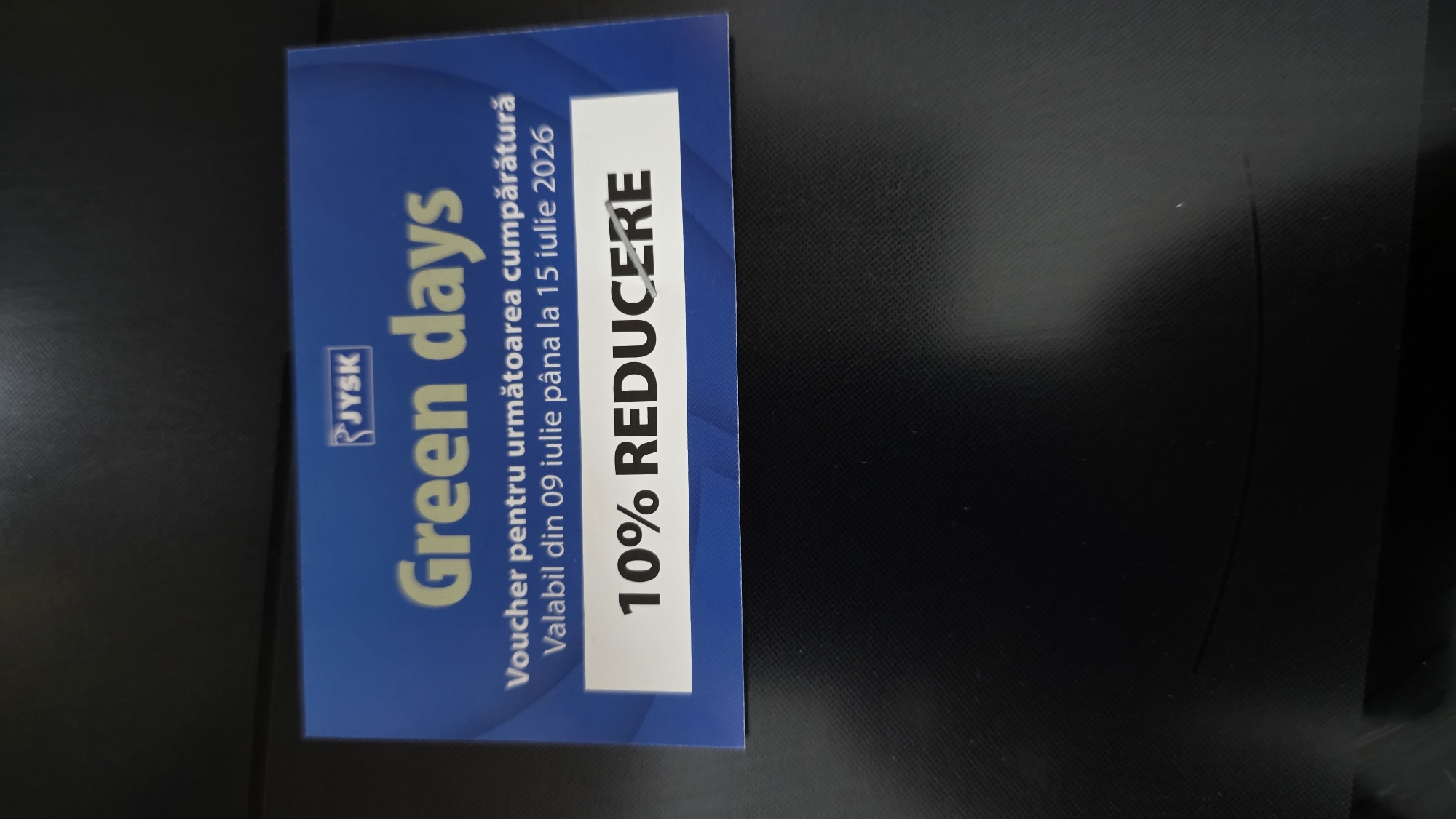 The image shows a close-up of a JYSK voucher. The voucher has a blue background with white text and a graphic element. The text reads "Green days" in large font. Below this, it says "Voucher pentru următoarea cumpărătură" which translates to "Voucher for the next purchase." Further down, it states "Valabil din 09 iulie până la 15 iulie 2026," meaning "Valid from July 9 to July 15, 2026." At the bottom, in a white rectangular box, is the text "10% REDUCERE," indicating "10% DISCOUNT." The voucher appears to be on a dark, textured surface, possibly a table or desk. There is no indication of any specific location in Chișinău, Moldova, nor are there any people or actions depicted.
