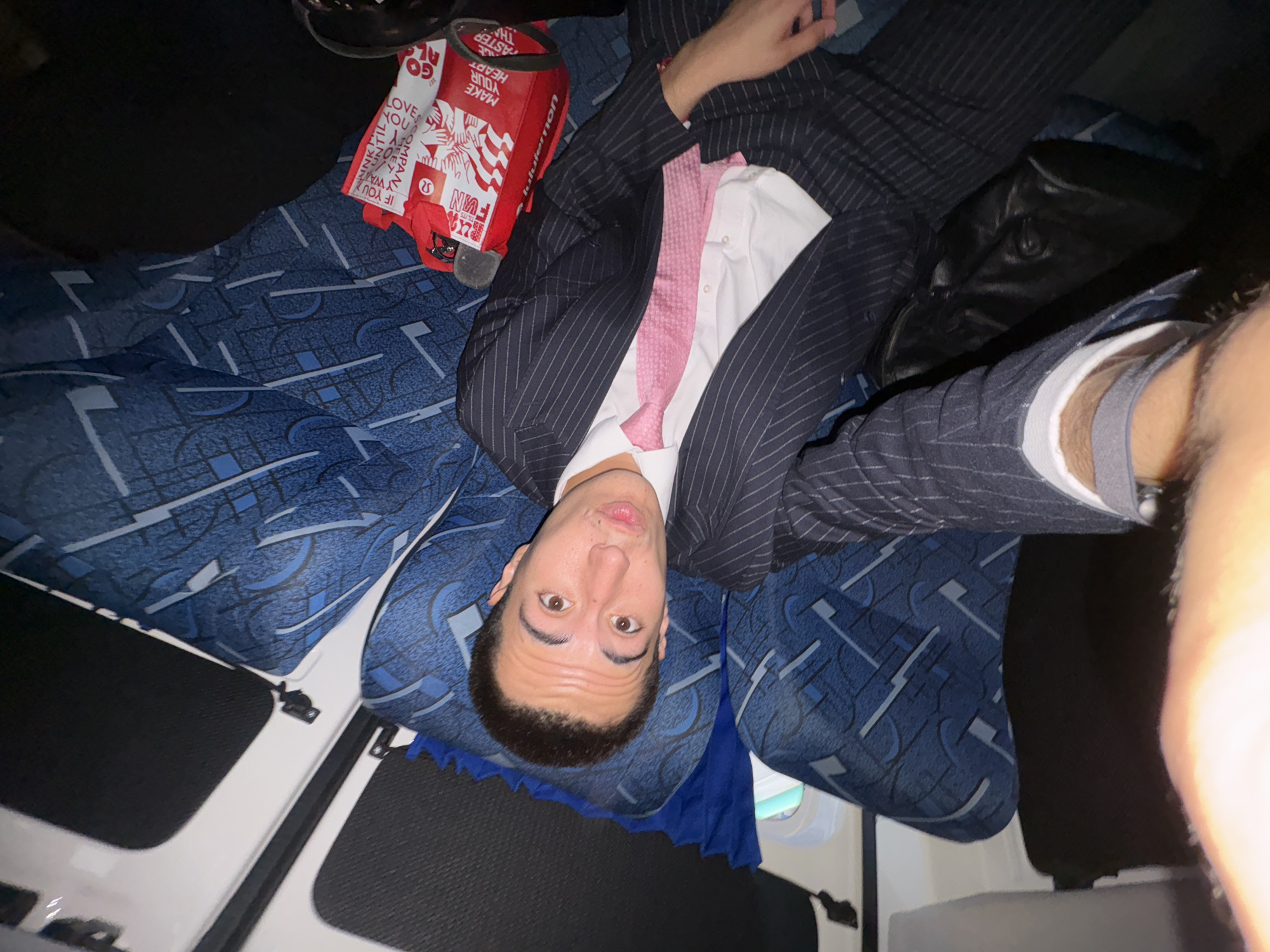 A man in a pinstripe suit and pink tie is lying in a bus seat. The bus has blue and white patterned upholstery. A red Lululemon tote bag sits next to him. He is taking a selfie, and part of his bare leg is visible. The bus window and interior are visible. The location is assumed to be in Mexico City, Mexico.