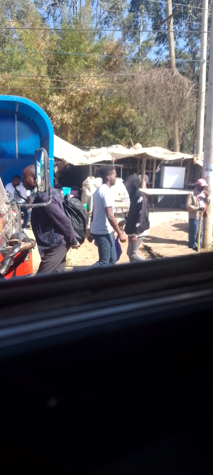 The image depicts a roadside scene in Ngong ward, Kenya, under clear daylight conditions. In the immediate foreground, a dark vehicle interior frame partially obscures the lower portion of the view. The mid-ground features a blue-colored vehicle, possibly a tuk-tuk or small utility truck, positioned on the left. A male subject, dressed in a dark jacket and black trousers, is partially visible from the back, holding a black backpack, standing adjacent to the blue vehicle. Another male subject with a bald head and white t-shirt is seated further left within or near the vehicle. To the right, two young male subjects are observed in close proximity; one wears a white t-shirt and blue jeans, holding a purple bag, while the other wears a black hooded jacket. Further right, a male subject in a light-colored shirt and hat, holding a wooden stick, stands near a light-colored utility pole. The ground surface consists of unpaved, exposed earth, with visible wooden planks forming a section of a path. In the background, several makeshift market stalls with white tarpaulin or corrugated metal roofs are present, one displaying a white refrigerator. Beyond these structures, dense green and dry vegetation, including tall trees and utility lines, are visible against a clear blue sky.