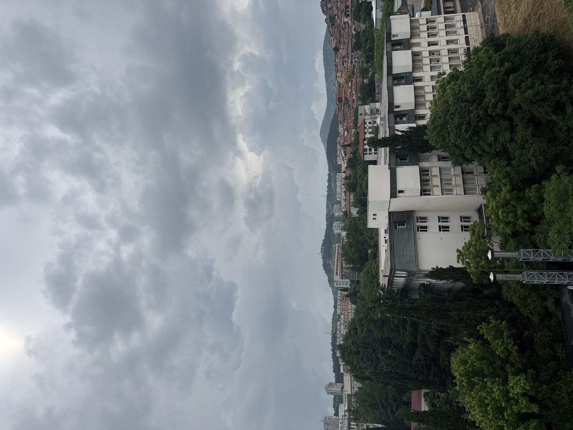 This is an outdoor, panoramic photograph of a city landscape in Stara Zagora, Bulgaria, taken under an overcast sky. The image is oriented vertically (rotated 90 degrees counter-clockwise), showing a significant portion of the sky at the top and the city sprawling below.
The sky is heavily clouded, with various shades of grey, indicating a cloudy day with diffused light. There are no clear signs of direct sunlight.
In the foreground and midground, a collection of multi-story buildings, predominantly white or light-colored, stand amidst numerous large, dense green trees. Some buildings feature dark, possibly flat or low-sloping roofs, and many visible windows. One particular building has a distinctive dark grey/green roof. Adjacent to some of these structures, a tall metal framework, resembling an antenna array or a support for lights, is visible.
Beyond these immediate structures, the cityscape extends across undulating hillsides. This part of the city displays a denser pattern of residential buildings, many characterized by traditional reddish-brown tiled roofs. The distant background reveals dark green, forested hills or mountains, completing the urban and natural panorama.
No specific human activity is visible, and the scene is static. There is no discernible text within the image. The overall impression is of a vast urban area nestled within a natural, hilly environment on a cloudy day.