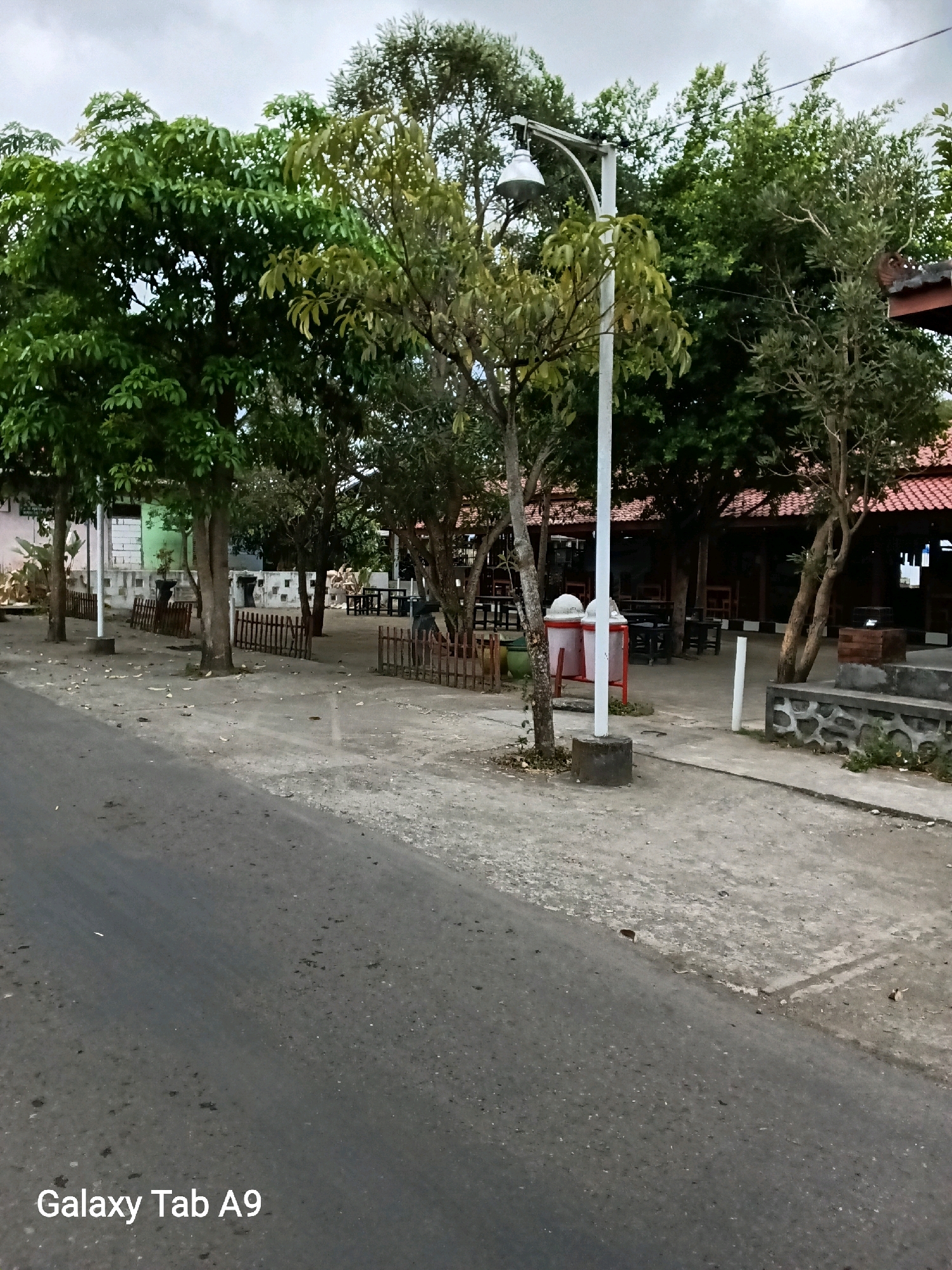 A medium shot captures a street scene in Kelutan, Indonesia, on a cloudy day. The foreground features an asphalt road with scattered debris. To the right, a paved walkway runs alongside the road, separated by a low, stone-lined planter. Beyond the walkway, a paved area contains several trees, two prominent trash receptacles on a red frame, and a lamppost. Further back, a building with a red tiled roof is visible, supported by wooden posts and featuring an open-air dining area with tables and chairs. A low wooden fence borders the paved area, and various plants and smaller trees are interspersed throughout the scene. The background is filled with lush green trees. The "Galaxy Tab A9" watermark is present in the bottom left corner of the image.