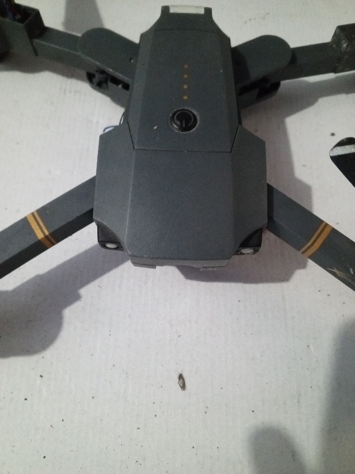 A gray drone sits on a textured white surface. The body of the drone is in the center, featuring a power button and battery indicator lights. Two arms with gold stripes extend towards the upper left and lower right corners. The shadows suggest the light source is above and slightly to the right. Debris is scattered on the white surface, including a dark speck near the bottom center.