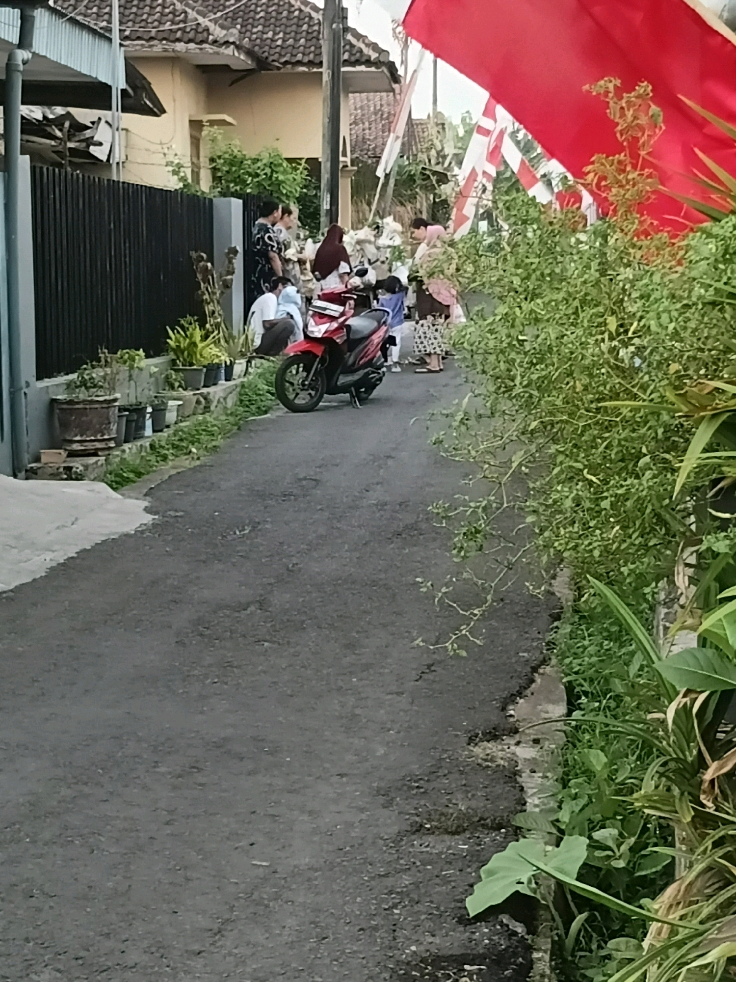 A narrow, paved street in the city of Kebonsari, Indonesia, is lined with green foliage and a black metal fence. Several people are standing near the fence and a red motorcycle is parked on the street. The people appear to be gathered for an event, possibly a celebration, as indicated by the Indonesian flags displayed prominently in the background. The houses in the background are simple, with tiled roofs. The scene suggests a residential area within the city.