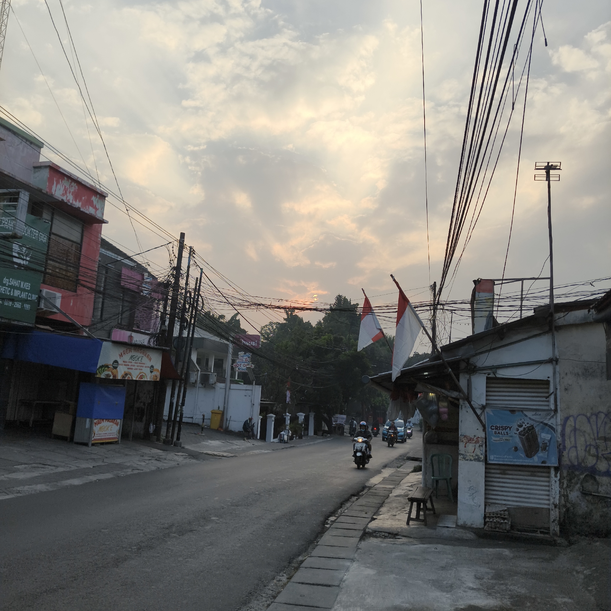 This image depicts a street scene in South Jakarta, Indonesia, at sunset. The sky is filled with clouds, illuminated by the setting sun, which is partially visible above a line of trees. The foreground shows a paved road with a sidewalk to the right. On the left side of the road, there are buildings with storefronts. One building has signs that read "drg. SAHAT M.KES ESTHETIC & IMPLANT CLINIC" and another advertisement for "MAEM MAKAN MBOKE". On the right side of the road, there is a smaller building with a "CRISPY BALLS" ice cream advertisement. Indonesian flags are visible on poles attached to the building on the right. Several motorcycles and a car are on the road, suggesting traffic. Overhead, a dense network of electrical wires crisscrosses the sky, typical of urban environments. The overall impression is a busy, lived-in street with a mix of commercial establishments and residential structures.