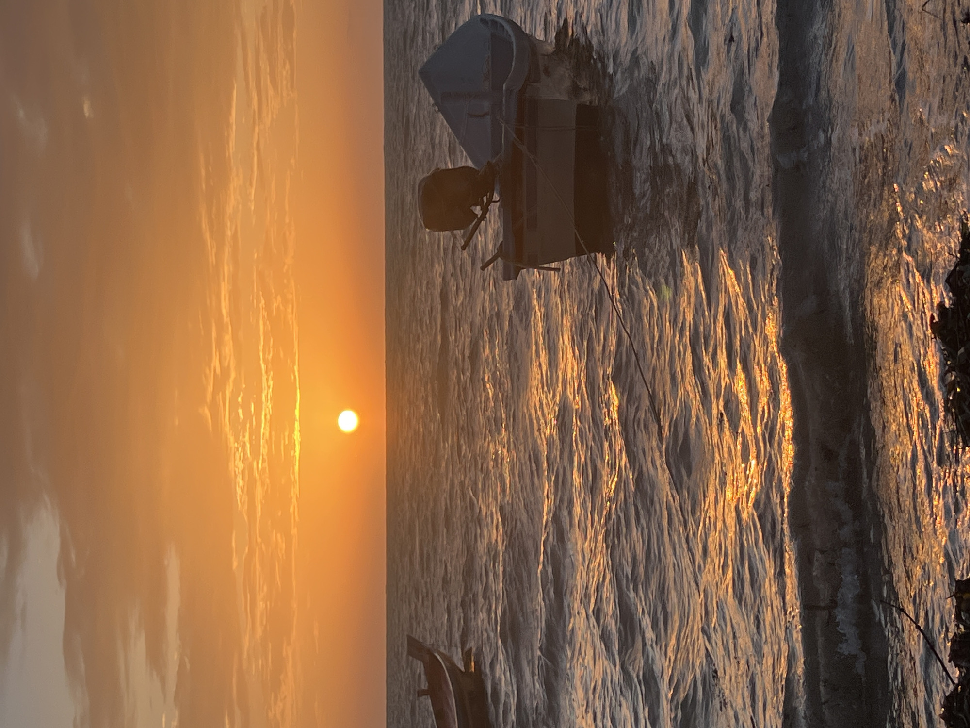 A vertical, medium shot captures a serene sunset over the waters of Ko Pha-ngan District, Thailand. The sun, a bright, warm orb, hangs low on the horizon, casting a golden glow across the sky and water. Wispy clouds, tinged with orange and pink, drift across the sky, adding texture and depth. The sea is choppy, with gentle waves reflecting the sunlight, creating a shimmering effect.
In the foreground, two small fishing boats are anchored. The boat on the right is closer to the viewer, its hull a muted blue and white, with a dark outboard motor mounted at the stern. A light blue tarp covers part of the boat. A thin rope extends from this boat towards the left of the frame. To the left, another boat is partially visible, its dark red hull contrasting with the muted tones of the water. The boats appear empty, suggesting a peaceful stillness to the scene.
The shoreline in the bottom foreground is dark and indistinct, appearing to be made of rocks and debris, with some dark plant matter. The overall atmosphere is tranquil and picturesque, evoking a sense of calm and natural beauty.
