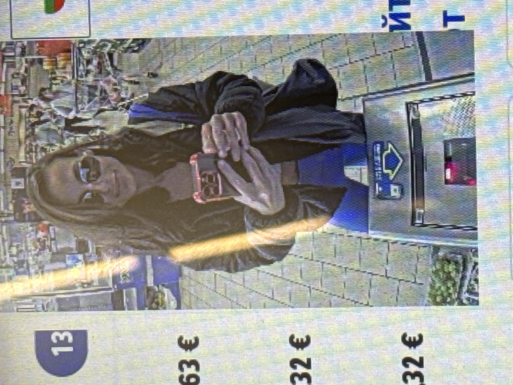 A woman wearing sunglasses, a blue top, and a black jacket is taking a selfie with a pink cell phone in Pleven, Bulgaria, next to a vending machine. Text above the vending machine is in Cyrillic. A price tag for €13.63 is visible in the bottom left of the frame. The scene appears to be outdoors, possibly on a sidewalk or in a public space.