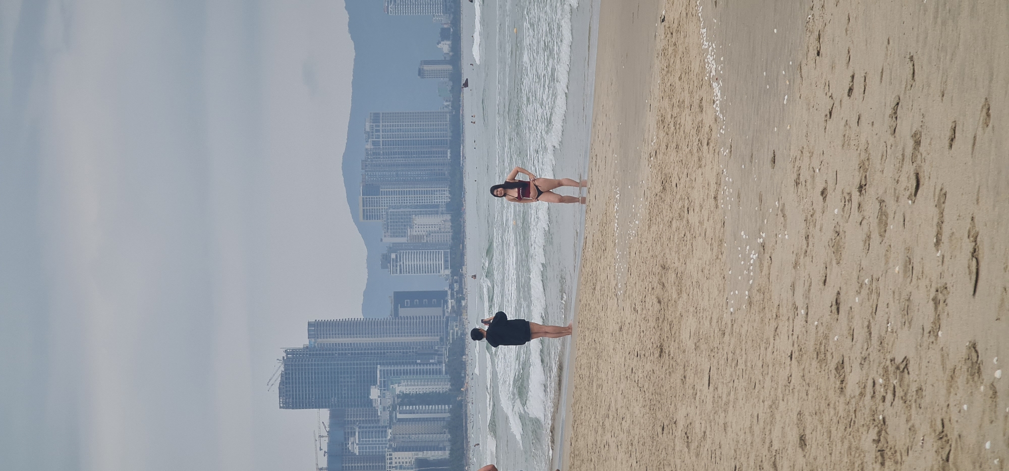 The image shows a beach in Đà Nẵng, Vietnam. In the background, there are tall buildings and a mountain range obscured by a light haze. Two individuals are standing on the beach. One person, wearing a black shirt and shorts, is holding a phone, appearing to take a picture. A second person, wearing a bikini, is posing near the water's edge. The sand is light brown and there are scattered white objects on the sand. Waves are breaking on the shore.