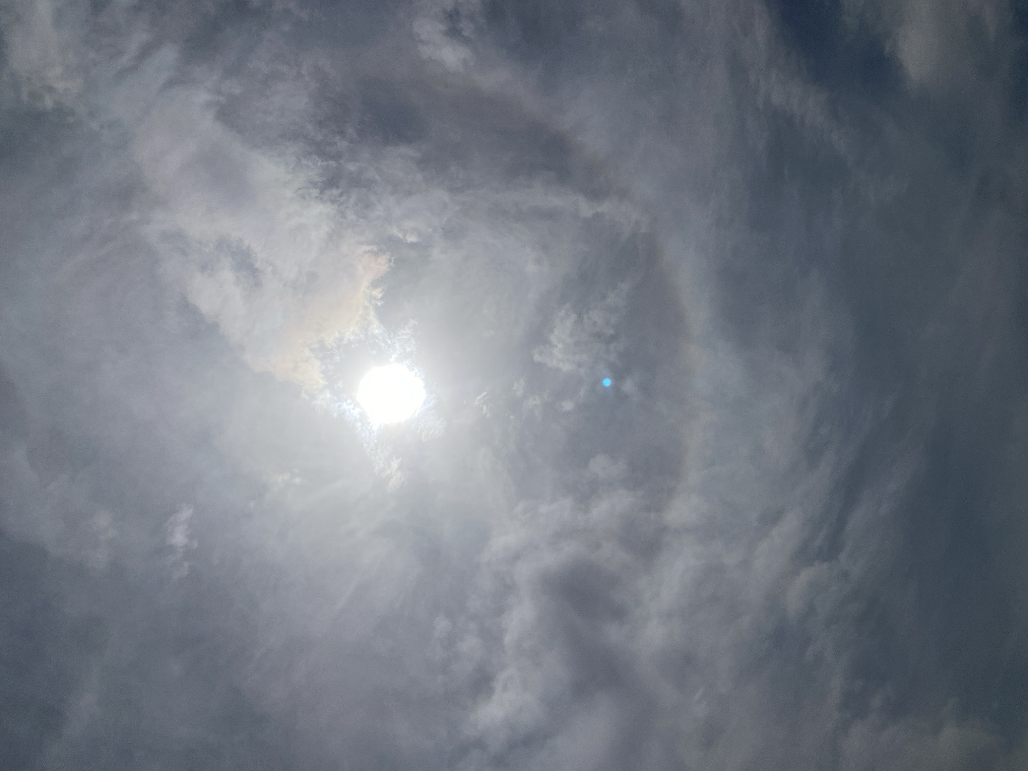 The image depicts a bright sun high in a cloudy sky above Barden Yana, Nigeria. The sun, a brilliant white orb, is positioned in the upper center of the frame, partially obscured by thin, wispy clouds that create a bright glare. Surrounding the sun, a distinct, faint circular solar halo, characterized by a subtle rainbow effect, is visible against the darker, predominantly gray and white scattered clouds that fill the rest of the sky. A small, prominent blue lens flare is present directly below the sun's position. No other subjects, objects, people, actions, or interactions are discernible; the scene is entirely an outdoor sky view.