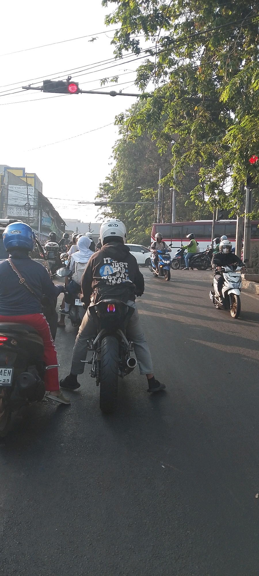 The image depicts a busy street scene in Bekasi, Indonesia, likely during the late afternoon due to the long shadows. The focus is on a group of motorcyclists stopped at a traffic light. A red traffic light is visible in the upper left corner, indicating a stop. In the foreground, a person wearing a blue helmet and a dark shirt is on a motorcycle, with red pants visible. To their right, another person in a white helmet and a black hoodie with the text "IT'S JUST A BAD DAY NOT A BAD LIFE" is also on a motorcycle. Several other motorcycles and riders are visible further down the street, navigating through traffic. A large red bus is seen in the background. The environment includes paved roads, overhead power lines, and lush green trees lining the street, suggesting an urban or semi-urban setting within Bekasi.