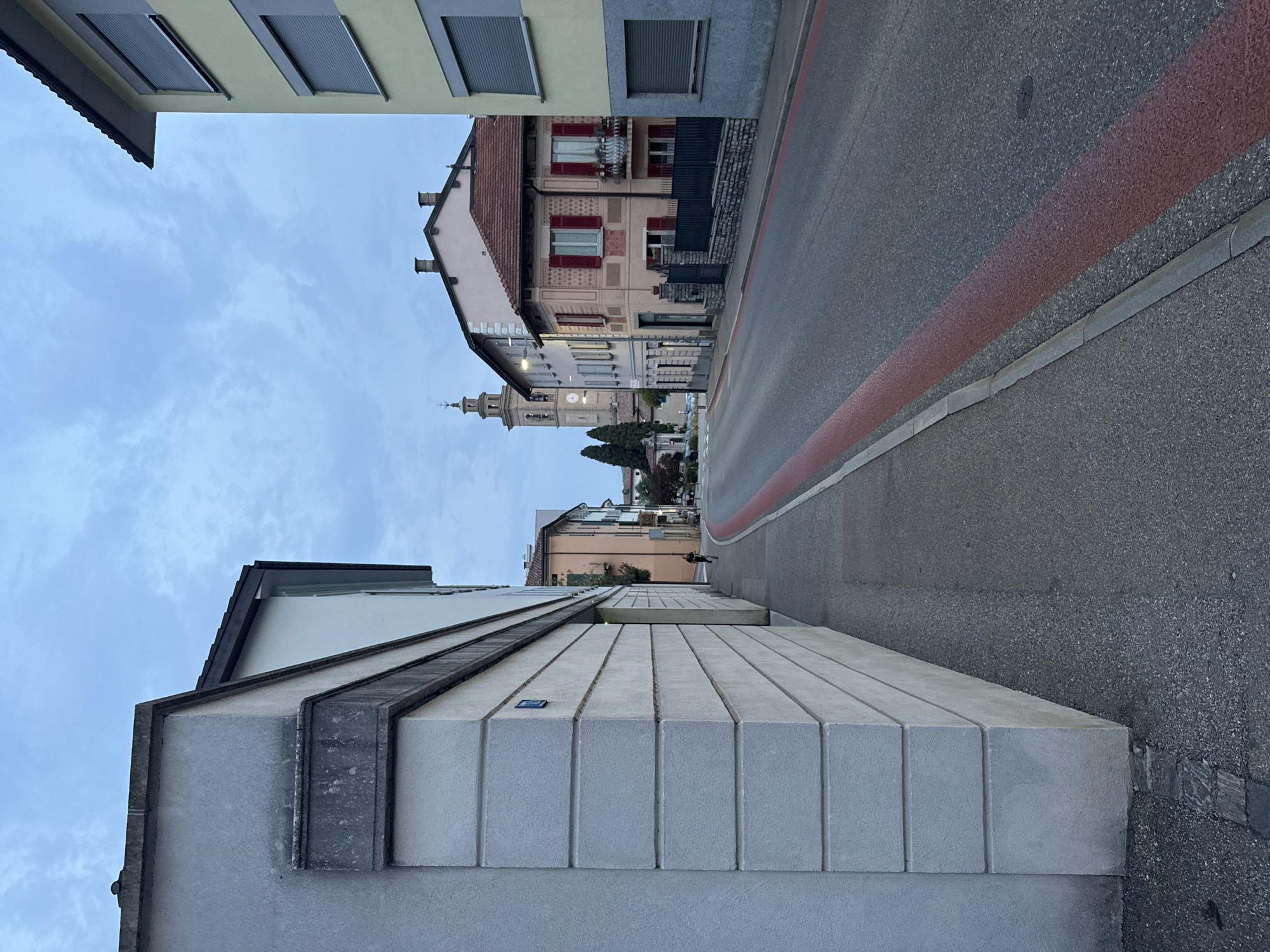 The image shows a street scene in Balerna, Switzerland, at dusk. A paved road runs diagonally from the bottom right to the top left of the frame, marked with a continuous red line. A sidewalk runs along the left side of the road, adjacent to a modern building with a textured grey facade. Further down the street, buildings of various architectural styles are visible, including a multi-story building with a bell tower. A single person is seen jogging on the sidewalk in the middle distance. The sky is overcast with a pale blue hue, indicative of twilight.