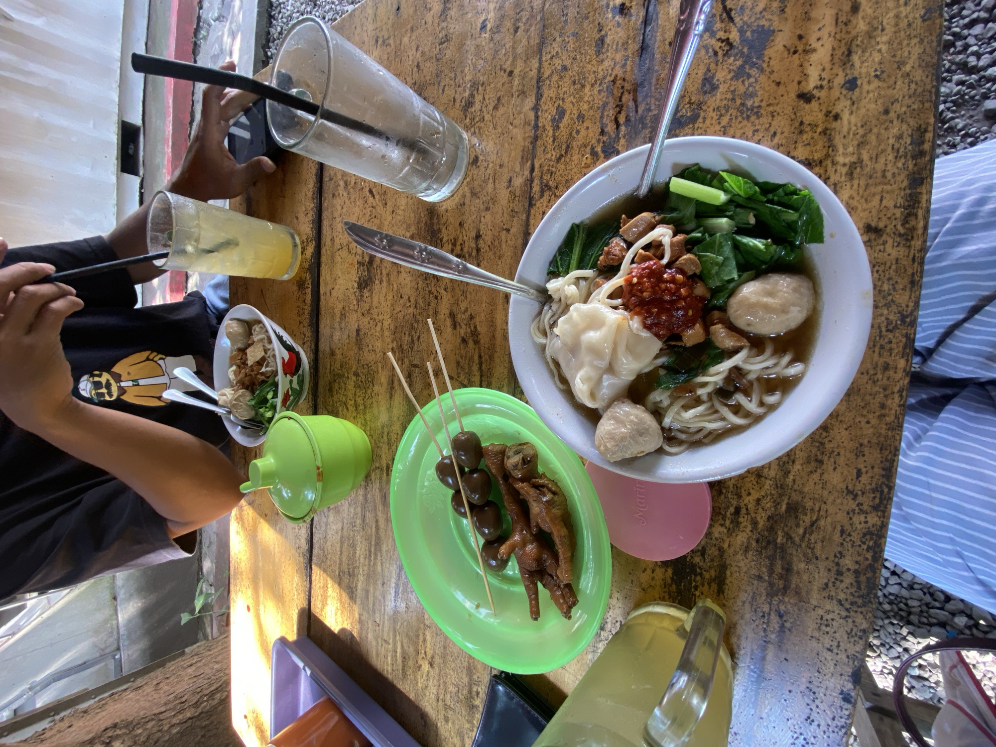 This media file captures a top-down view of a casual meal set on a rustic wooden table, likely at an outdoor or semi-outdoor eatery in Madiun, Indonesia.
The primary subject is a large bowl of *Mie Ayam Bakso* (chicken noodles with meatballs) served in a white bowl, positioned slightly to the right of the center. It contains noodles, dark brown chicken pieces, green leafy vegetables (possibly bok choy), two large meatballs, and a *wonton* or *pangsit* dumpling, topped with a dollop of red chili paste. To the left of the main bowl, a bright green oval plate holds skewers of *sate ceker* (chicken feet) and *sate telur puyuh* (quail eggs), indicating common Indonesian street food snacks. A smaller bowl of noodles is also visible on the far left.
Beverages include a clear glass with a yellow/orange drink and a black straw, held by a person on the left whose hands and part of a black t-shirt with a yellow graphic are visible. Another empty clear glass with a black straw is near the top center. A yellow jug or pitcher is seen in the bottom right corner.
Two individuals are partially visible. On the left, a person is holding a drink and appears to be interacting with a phone. On the right, only a leg wearing light blue and white striped pants is visible.
The wooden table has a worn, light brown appearance. Various items are scattered across it, including spoons, a green lidded condiment container, a black wallet, and a pink round coaster with the word "Marin" faintly visible. The background shows a light-colored wall with red accents on the left and a gravel ground area on the right, suggesting a bright, sunny daytime setting. The scene evokes a relaxed, informal dining experience.