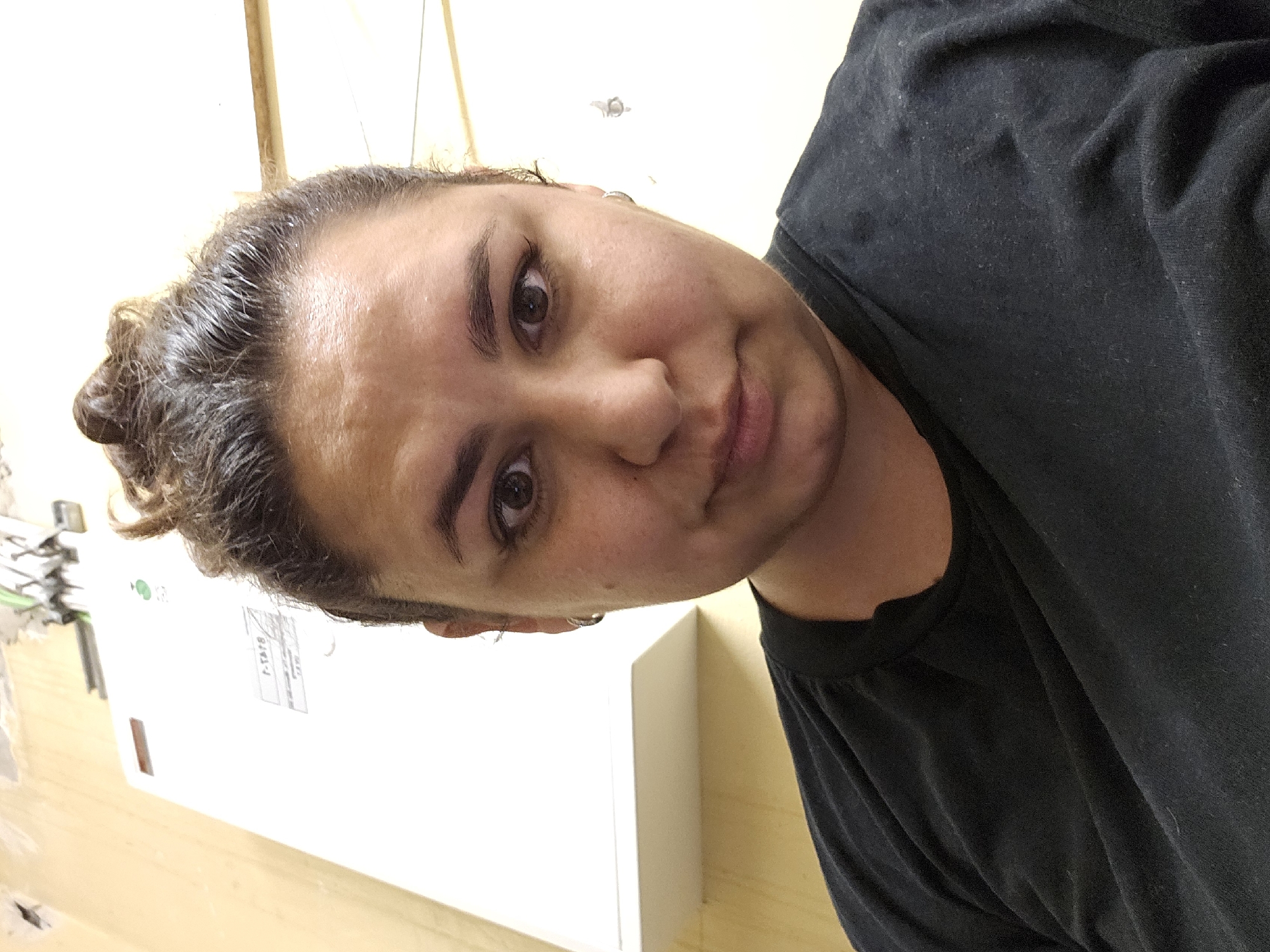 A selfie of a woman with dark, curly hair pulled up into a bun is shown. She is wearing a black t-shirt. Her face is partially obscured by a white rectangular object in the background, which appears to be a piece of equipment with some labels. A beige wall is visible to the left, and a white wall to the right. The scene appears to be indoors, possibly in a utility area or workshop. The lighting is artificial. The image is shot from a close-up perspective, focusing on the woman's face and upper torso. The background suggests an industrial or commercial setting.