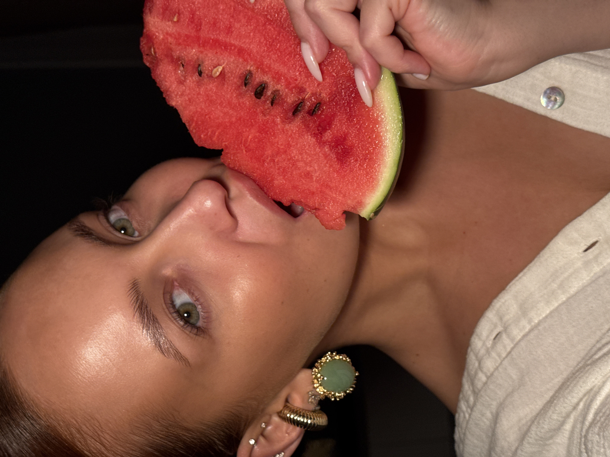 An overhead close-up shot captures a person biting into a large slice of fresh red watermelon. The person, likely female, has smooth, lightly tanned skin, striking green eyes, and well-defined eyebrows. Her mouth is slightly open, positioned to eat the watermelon, which appears juicy with visible black seeds embedded in the flesh and a thin green rind.
A hand with long, light-colored, pointed fingernails holds the watermelon. The person is wearing a white or off-white textured shirt, partially visible, with a mother-of-pearl button on the collar. Her right ear is adorned with several piercings: a prominent large, circular, gold-rimmed earring with a green stone (possibly jade), a gold ribbed ear cuff on the cartilage, and multiple small stud piercings.
The background is predominantly dark, suggesting an indoor setting or nighttime. The lighting is dramatic, illuminating the person's face and the watermelon, creating highlights on her skin and the fruit's wet surface. The image is tilted, providing a dynamic and intimate perspective on the act of eating the watermelon. There is no visible text. The activity takes place in what could be a relaxed setting, possibly in Chaikovsky, Russia, based on the provided context.