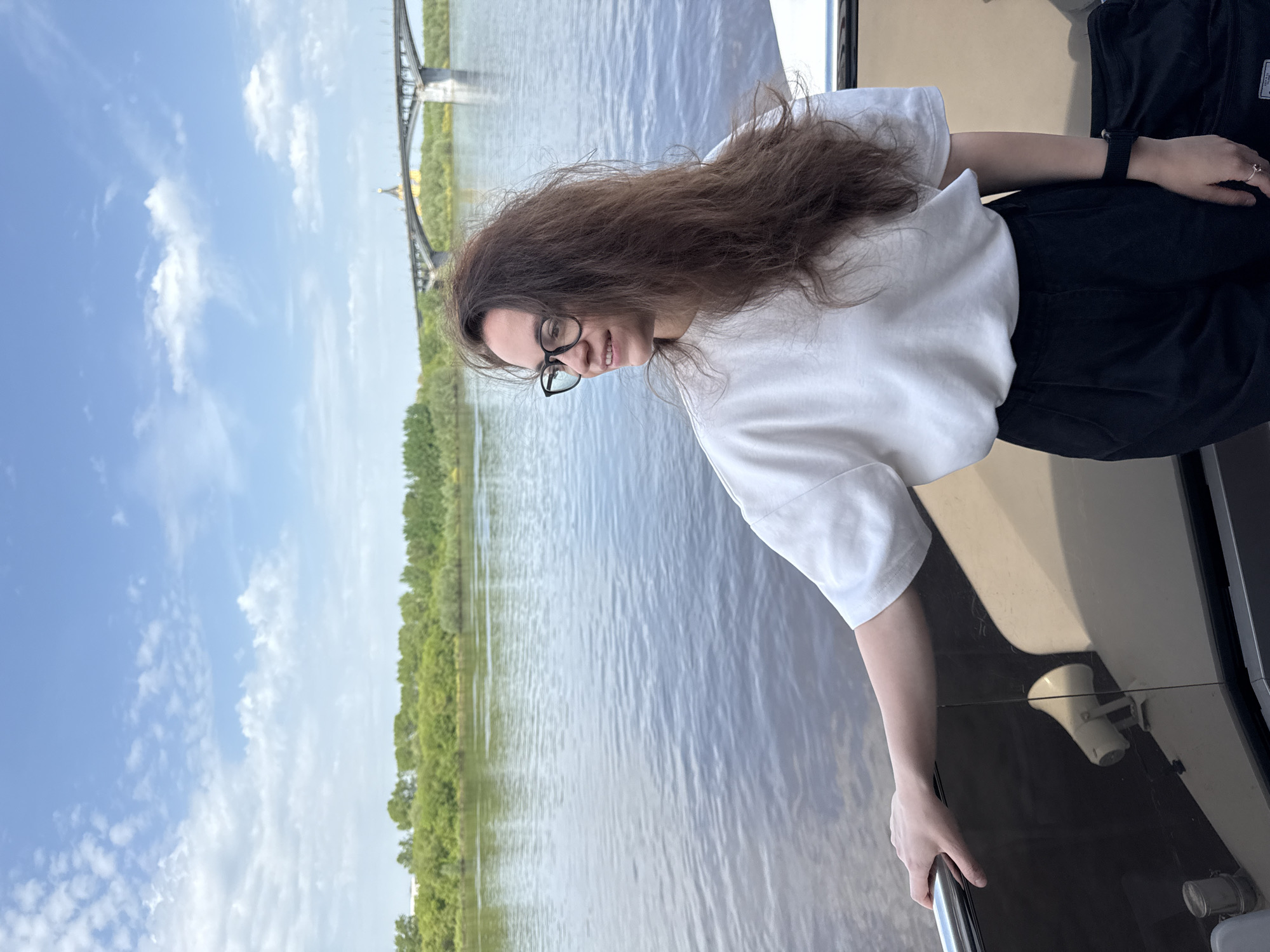 A medium shot depicts a woman on a boat, with a cityscape including a bridge visible in the background. The woman has long, brown, wavy hair and is wearing glasses, a white t-shirt, and dark pants. She is holding onto a railing on the boat. The environment includes a river, trees lining the riverbank, a blue sky with clouds, and a bridge in the distance. The location is Nizhny Novgorod, Russia, as indicated by the distinctive bridge architecture.