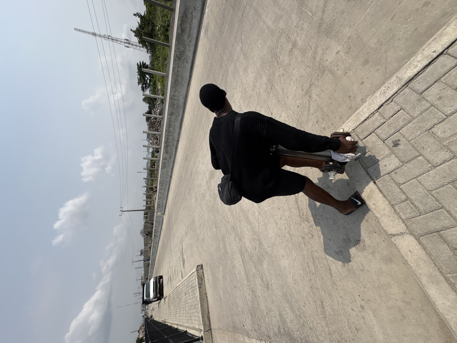 The image captures an outdoor scene in Igboefon, Nigeria, under a bright, partly cloudy sky. A dark-skinned individual, seen from behind and bending forward, is actively engaging with a metallic lifting device, possibly a hydraulic jack, positioned at the juncture of a light gray concrete roadway and a darker gray brick-paved sidewalk. The individual wears a black ribbed long-sleeved top, black shorts, black Nike slides on their feet, and a black head covering; a black cross-body bag is slung over their back. Their right hand, wrapped in a white cloth, grips the device's handle, which exhibits metallic disc-like components near its base.
The concrete roadway extends into the background, flanked on the left by a low concrete curb and a black metal fence bordering the sidewalk. A white pickup truck is discernible in the far distance on the left side of the road. To the right of the roadway, a concrete barrier delineates an area characterized by scattered construction materials, rudimentary fencing, sparse vegetation, and several utility poles supporting overhead power lines. A tall, lattice-structured communication tower is visible in the far background on the right. Strong shadows indicate direct sunlight.