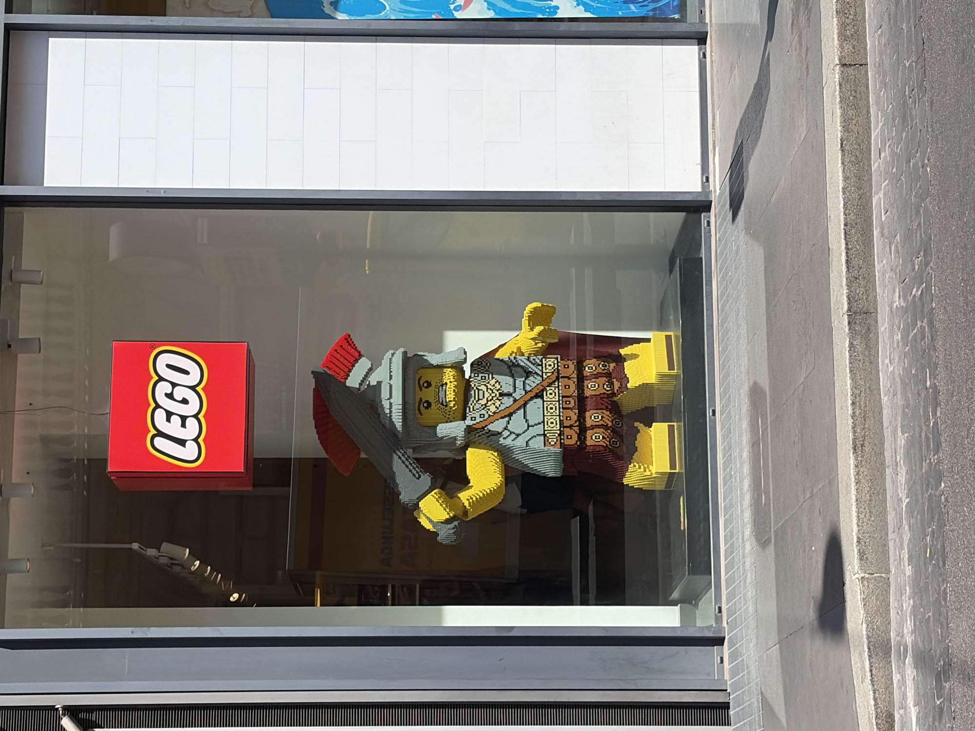 A full-shot, eye-level photograph captures the exterior of a LEGO store in Rome, Italy. The store features a large display window showcasing a life-sized LEGO Roman soldier. The soldier, constructed from yellow, gray, red, and brown LEGO bricks, stands prominently with a sword raised above its head. It wears a helmet, a tunic, armor, a cape, and a belt with a patterned design. The LEGO logo, a red square with "LEGO" written in white with a yellow border, is displayed on the upper left side of the window. The background of the store is visible through the glass, with some shelving and a blurred image of a person in the distance. The exterior of the store is framed by dark gray vertical beams and a white tiled wall on the right. The pavement in front of the store is paved with grey square tiles and a darker cobblestone street runs in front of it. Shadows of people and objects are cast on the pavement.