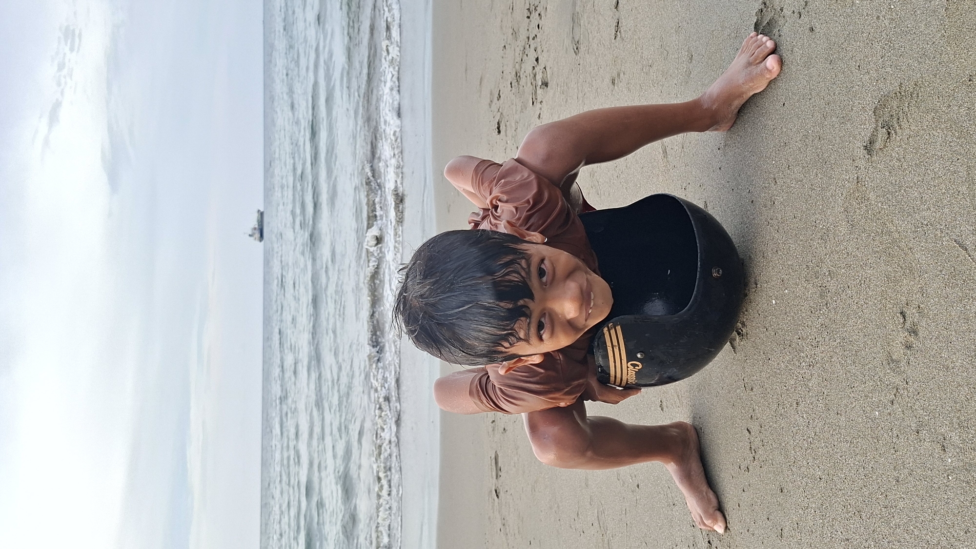 A young boy is on a sandy beach, positioned low to the ground, with his hands and feet supporting his weight. He is looking directly at the camera with a playful expression. In front of him, on the sand, is a black helmet with a gold stripe and the word "Classic" written on it. The ocean with gentle waves is behind him, and a large ship is visible in the distance on the horizon. The sky is overcast. The location is the beach in Lhokseumawe, Indonesia.