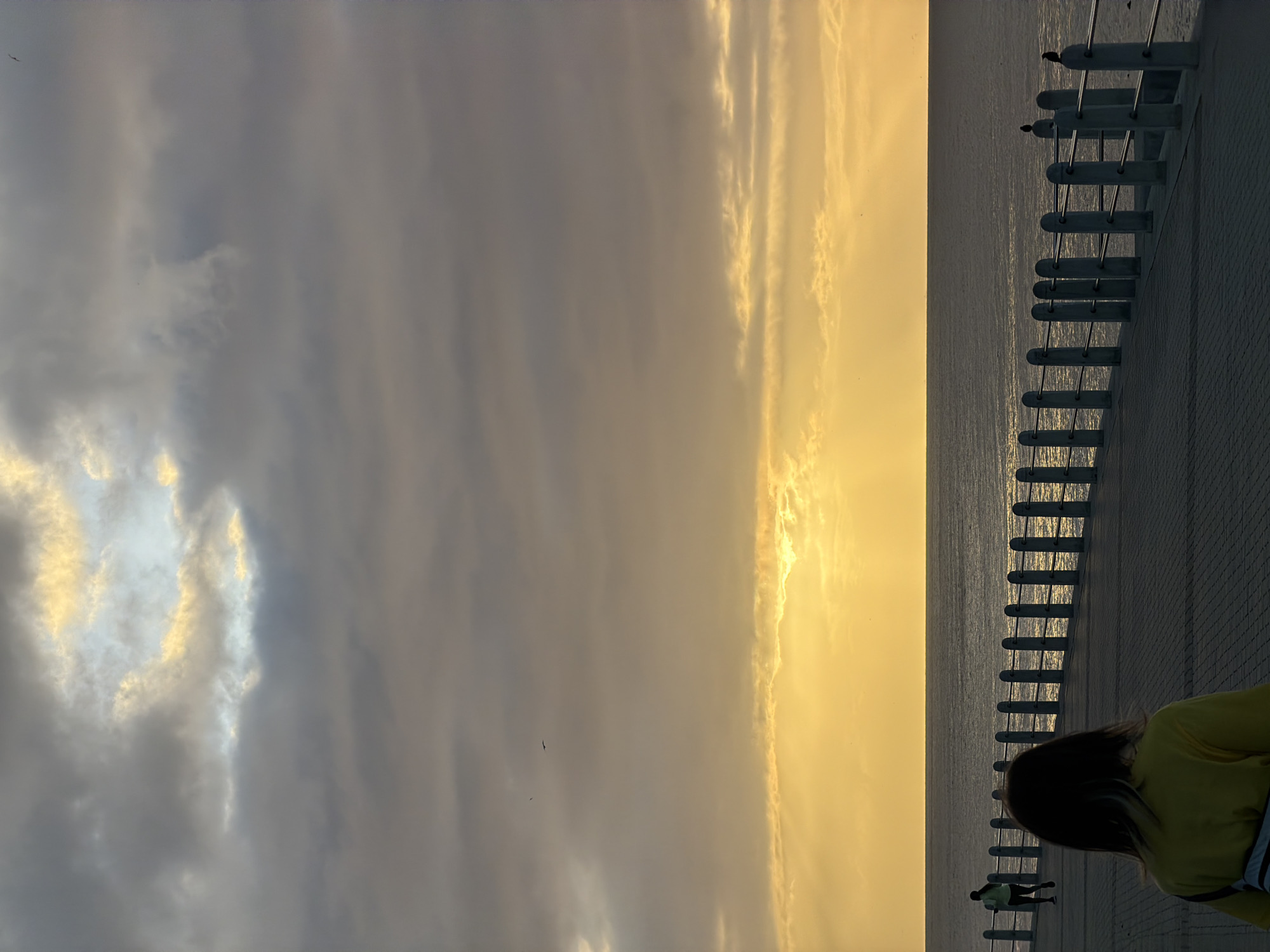 The image captures a scene at dusk or dawn in Cape Town, South Africa, along the coastline. The sky is a mix of gray and golden clouds, with the sun's light reflecting on the ocean. A concrete promenade runs along the coast, bordered by a series of evenly spaced concrete bollards connected by metal railings. A few people are present: one person wearing a yellow shirt is visible in the foreground, and two figures can be seen walking in the distance along the promenade. Two birds are perched on the railing near the horizon. The overall atmosphere is tranquil and serene, with a clear contrast between the city infrastructure and the natural environment.